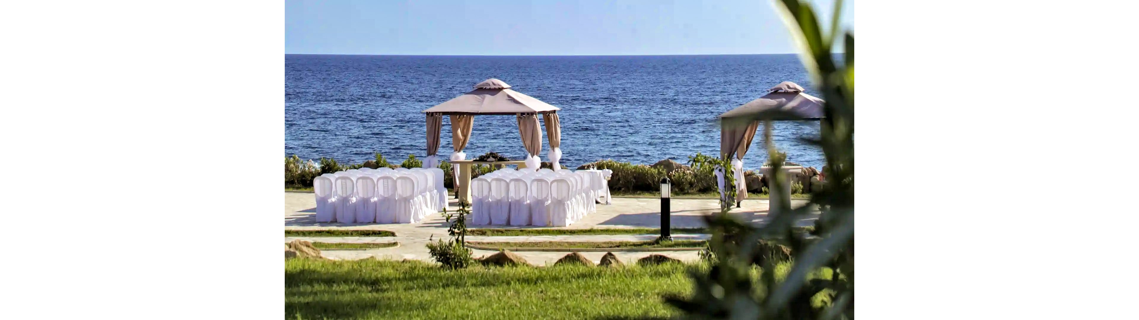 An elegant outdoor wedding ceremony setup is arranged beside a calm blue sea. Two fabric-draped gazebos frame rows of white-covered chairs facing the water. The scene is surrounded by green grass, low shrubs, and a paved path leading to the seating area. Bright daylight and clear skies create a serene, romantic atmosphere for a coastal event.