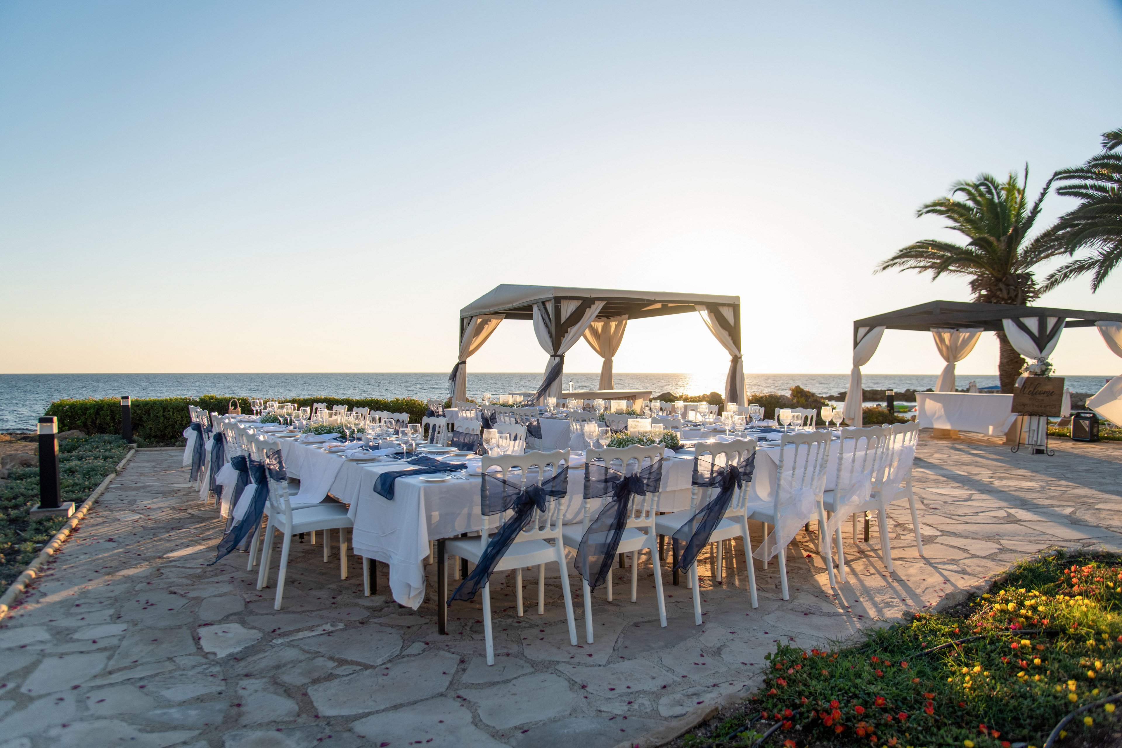 An elegant outdoor reception is arranged on a stone terrace overlooking the sea at sunset. Long banquet tables are covered with white linens and decorated with glassware, plates, and blue ribbon chair accents. Open-sided canopies frame the dining area, while a palm tree and low hedges border the space. The calm ocean horizon and warm evening light create a romantic, celebratory atmosphere.