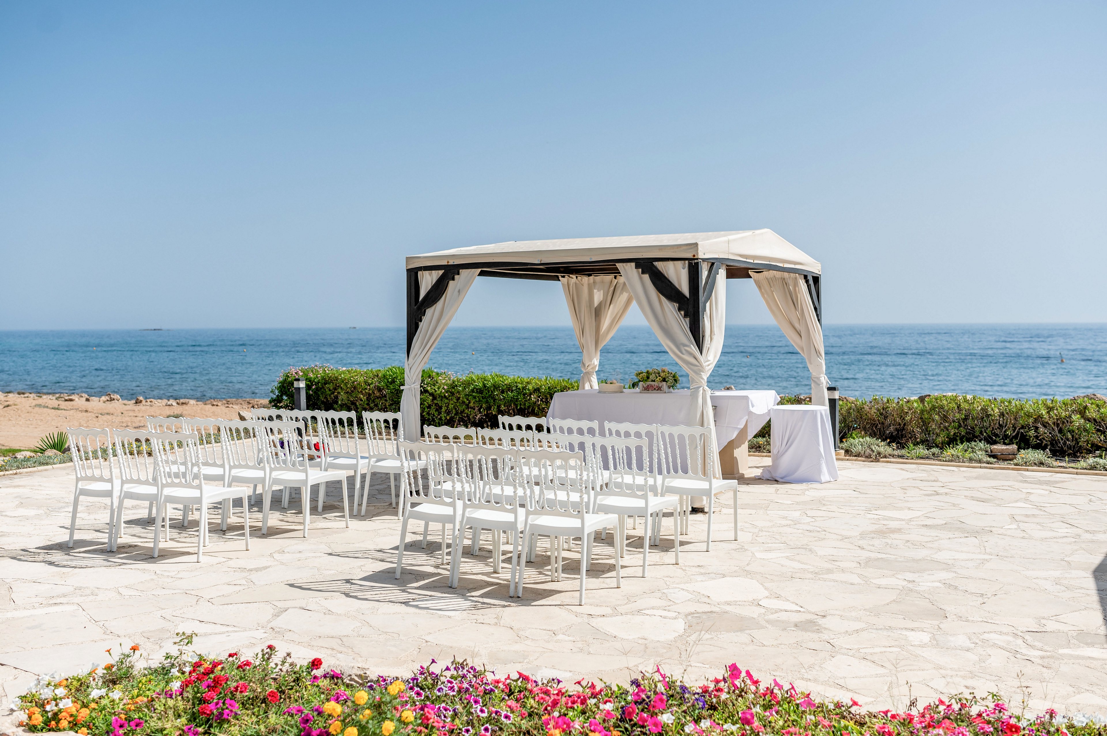 An elegant outdoor wedding ceremony setup is arranged on a stone terrace by the sea. A white gazebo with flowing curtains stands at the front, sheltering a small table and decor. Neatly aligned white chairs face the gazebo, creating an aisle for guests. Colorful flowers border the foreground, while the calm blue ocean and clear sky provide a serene backdrop.