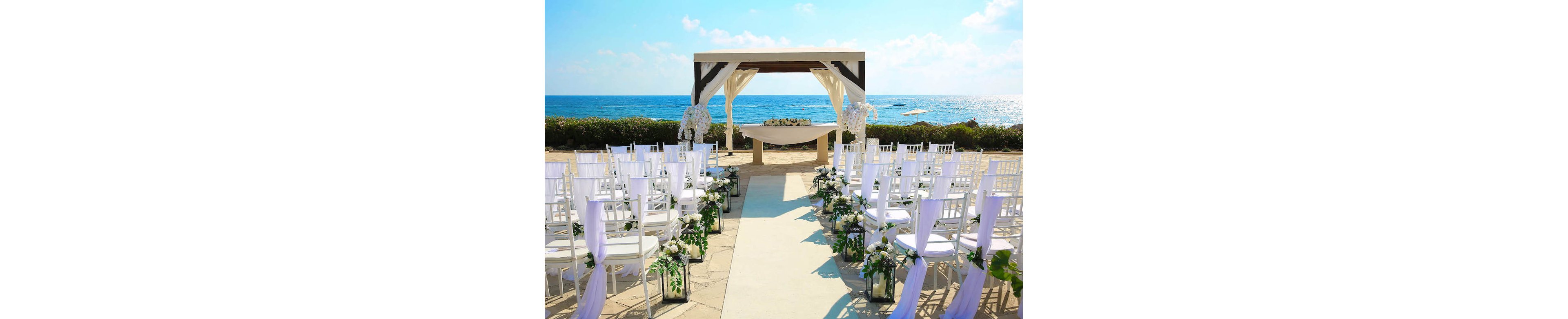 An outdoor wedding ceremony is arranged on a stone terrace overlooking a bright blue ocean. White chairs line both sides of an aisle leading to a draped wooden arch and altar table. Floral arrangements and greenery decorate the aisle and lanterns, creating a romantic atmosphere. The clear sky and calm sea enhance the light, airy mood of the scene.