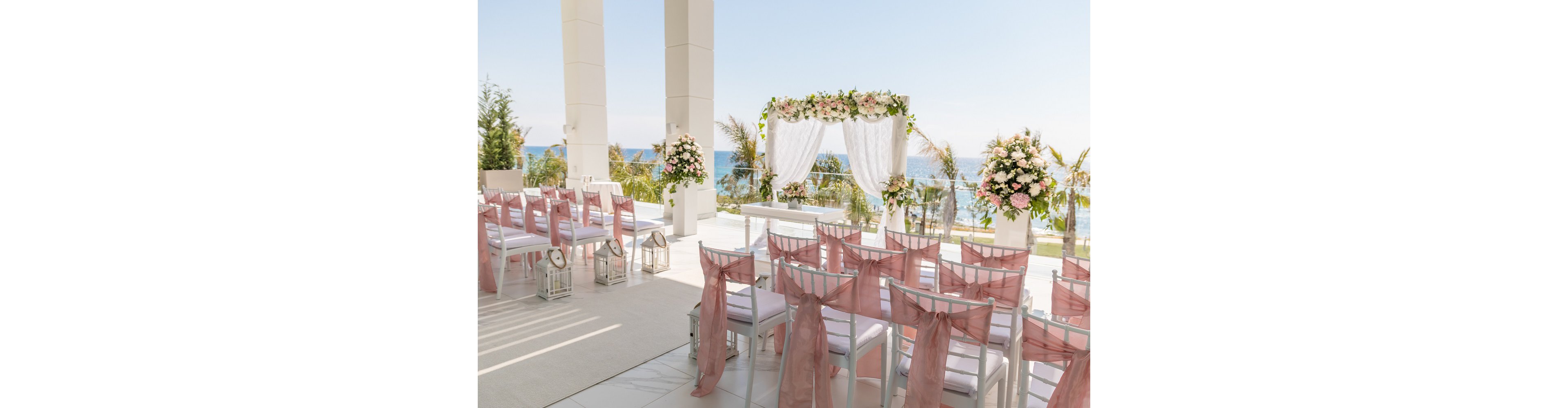 An outdoor wedding ceremony area is arranged on a bright terrace overlooking the sea. White chairs with soft pink fabric sashes are lined in rows facing a floral arch draped in white curtains. Large flower arrangements and decorative lanterns accent the aisle, creating a romantic and refined atmosphere. Palm trees and a clear blue sky complete the serene coastal backdrop.