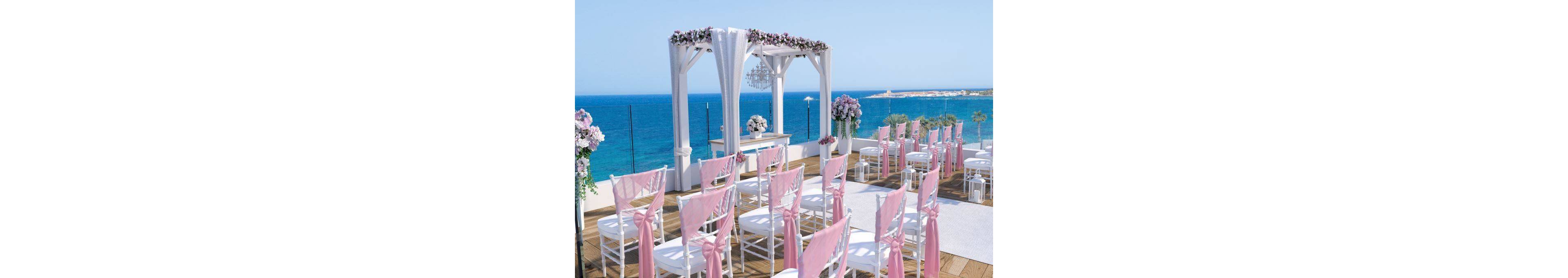 An elegant wedding ceremony area is arranged on a wooden deck overlooking a bright blue sea. White chairs with pink fabric sashes face a floral-decorated arch and altar table. Tall flower arrangements and a chandelier under the canopy add a luxurious touch. The clear sky and ocean horizon create a serene, romantic atmosphere for an outdoor celebration.