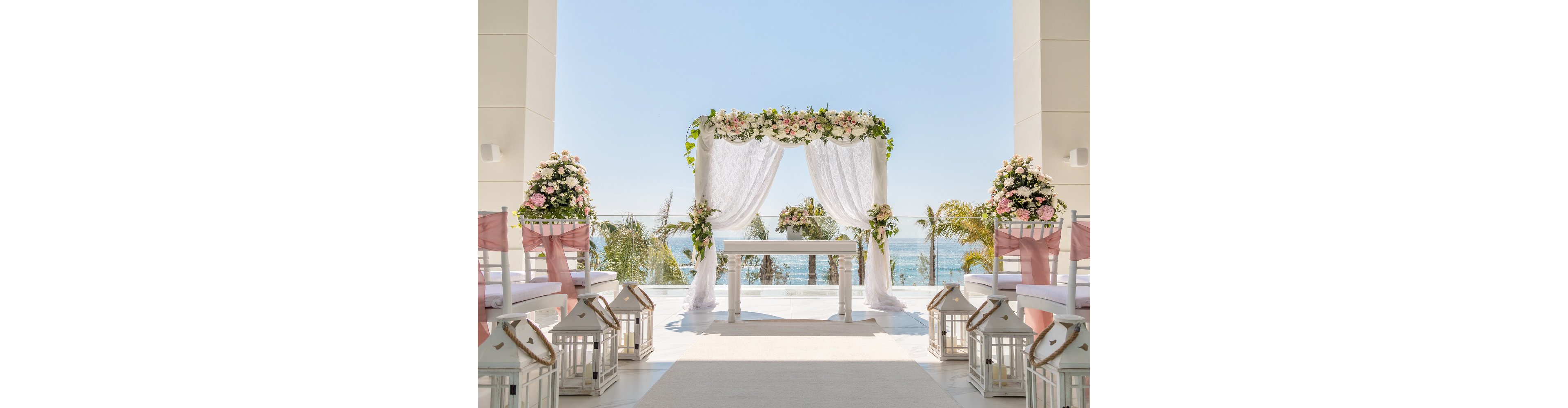 An elegant wedding ceremony setup features a white altar framed by a floral arch with pink and white flowers. Rows of white chairs decorated with soft pink sashes line the aisle, leading toward the ocean view in the background. White lanterns with candles are placed along the aisle, enhancing the romantic atmosphere. Palm trees and clear blue sky complete the bright, airy seaside setting.