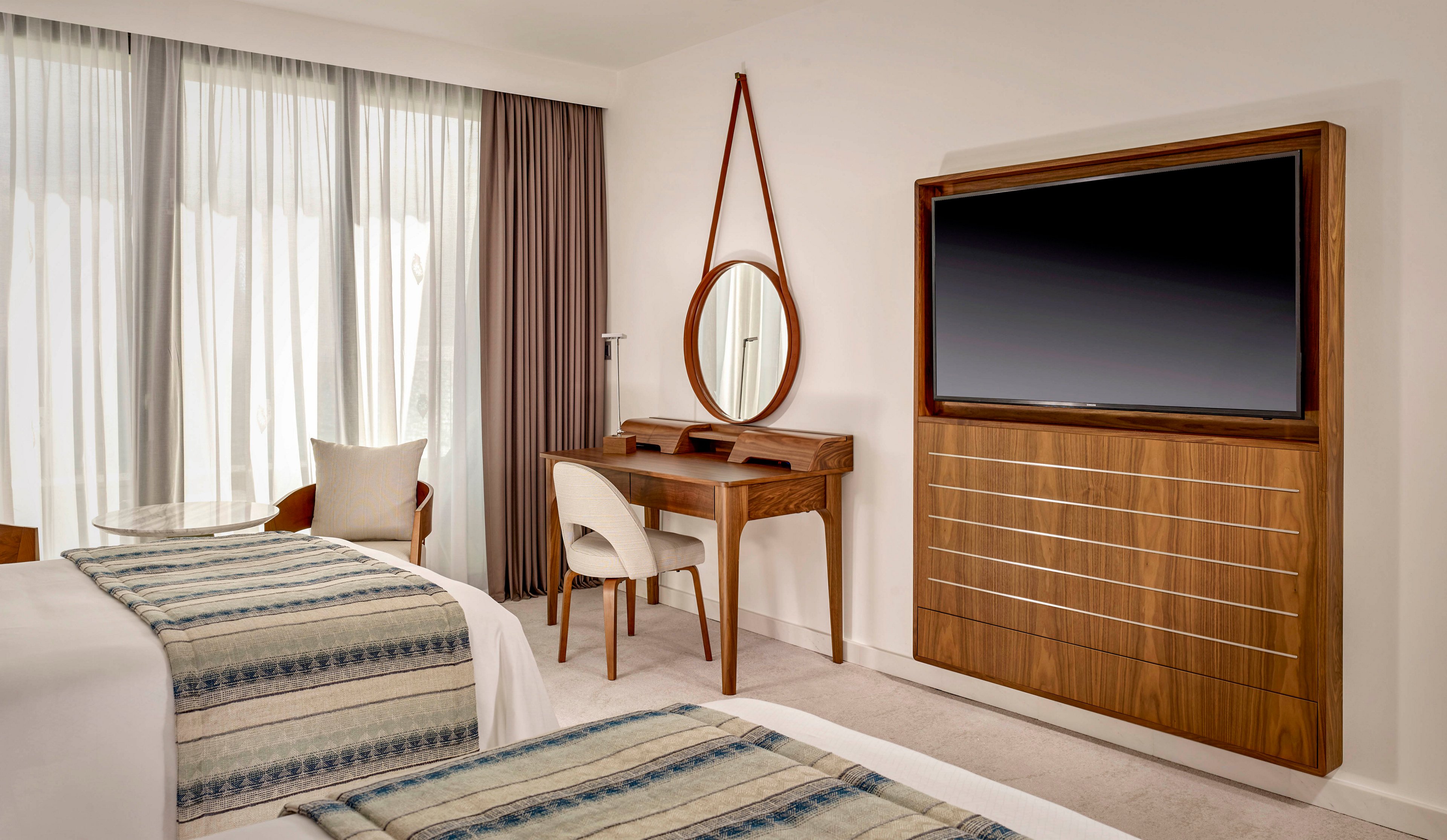 A contemporary hotel room features two neatly made twin beds with patterned throws. A large flat-screen television is mounted on a wooden cabinet opposite the beds. Beside it, a wooden vanity table with a round mirror and upholstered chair sits against the wall. Floor-to-ceiling windows with sheer curtains and drapes allow soft natural light to fill the minimalist, neutral-toned space.