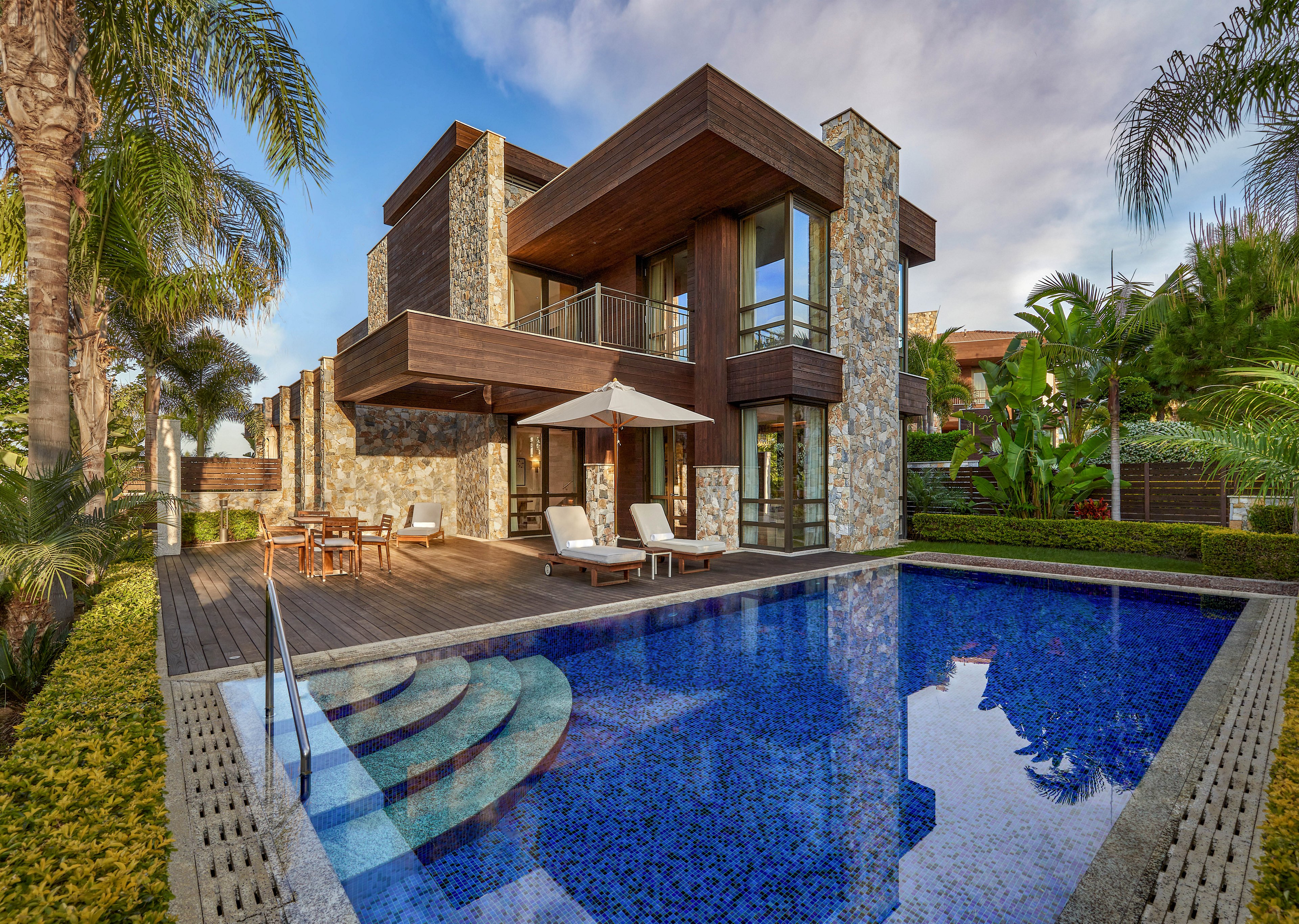 A contemporary two-story villa with stone and wood exterior overlooks a clear blue swimming pool. The scene features a spacious wooden deck with lounge chairs and a patio umbrella arranged for outdoor relaxation. Lush tropical landscaping, including palm trees and manicured hedges, surrounds the property. Warm sunlight and a partly cloudy sky enhance the tranquil, resort-like atmosphere.