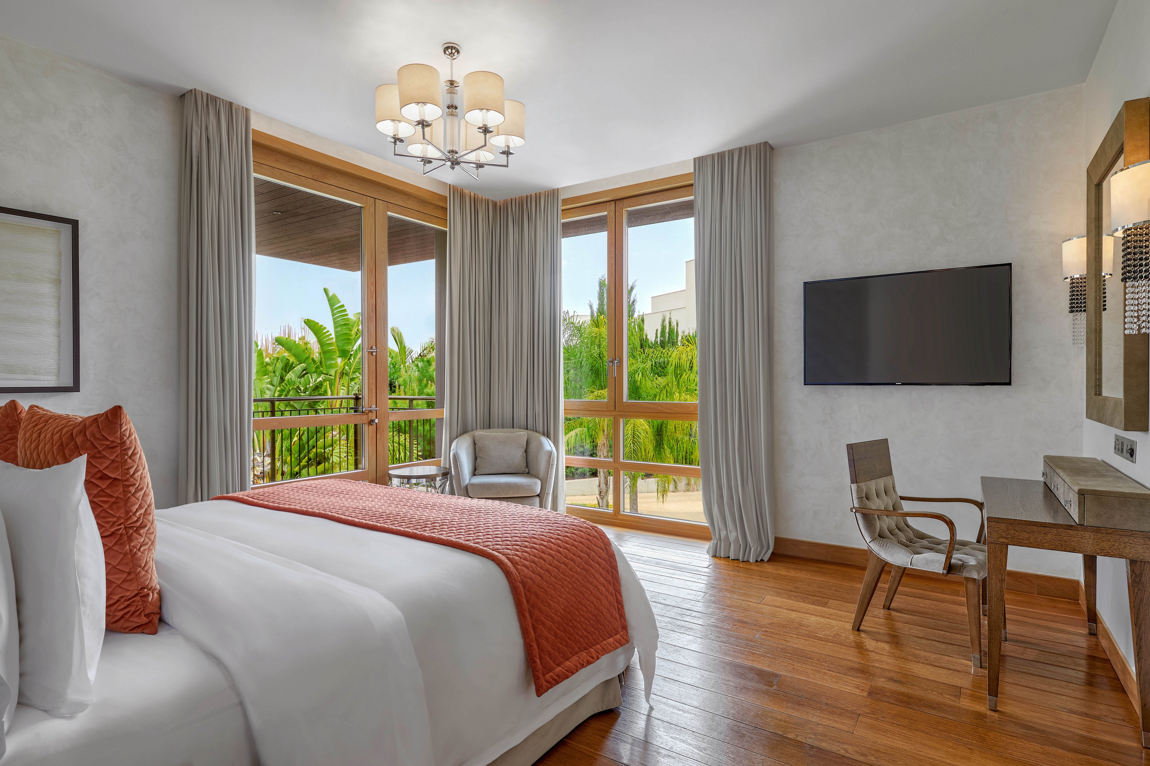 A contemporary hotel bedroom features a neatly made bed with white linens and an orange textured throw. Large floor-to-ceiling windows with light gray curtains open to a balcony overlooking lush green palm trees. A wall-mounted flat-screen television faces the bed, and a wooden desk with a chair sits to the side. Warm wooden flooring and soft neutral walls create a calm, inviting atmosphere.