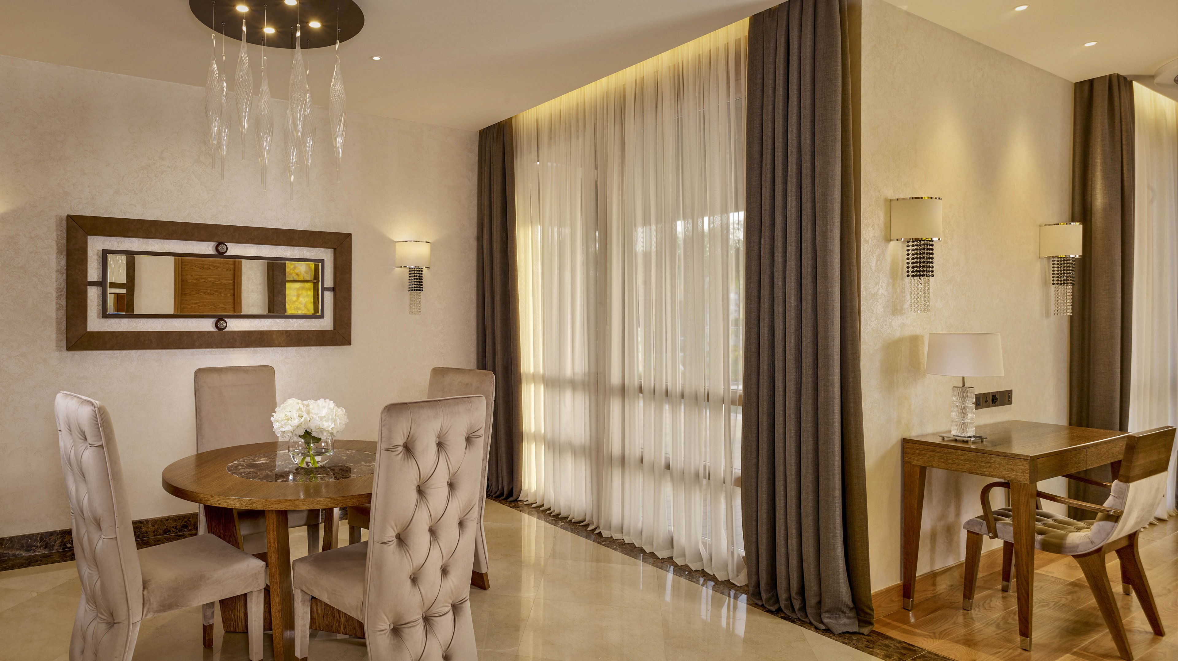 A sophisticated dining room features a round wooden table surrounded by upholstered chairs. Floor-to-ceiling sheer curtains with dark drapes cover large windows, allowing soft natural light into the space. A modern chandelier with hanging glass elements and wall sconces provide additional ambient lighting. A small wooden desk with a chair sits against the wall, creating a combined dining and work area in a neutral, contemporary interior.