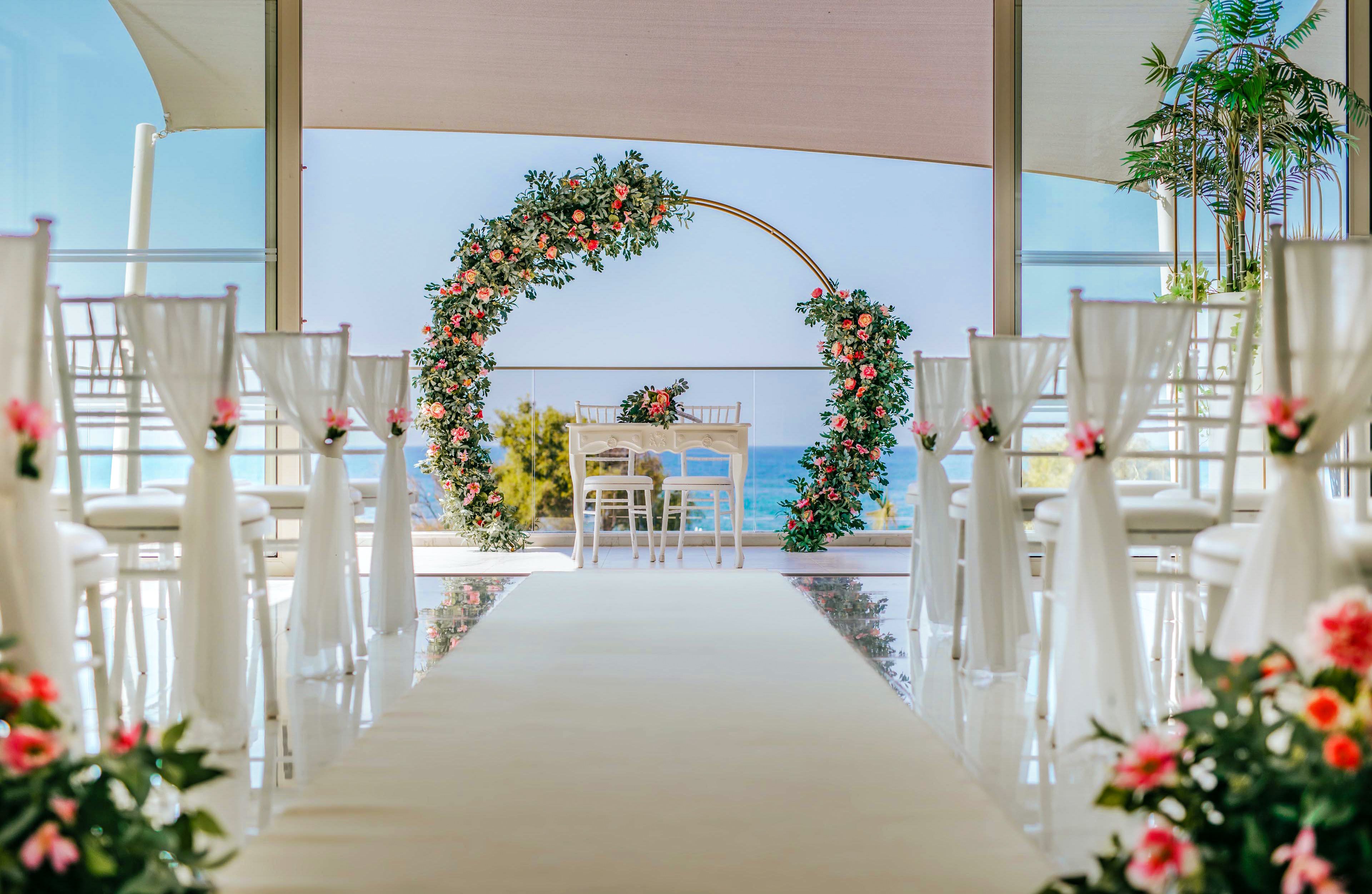An elegant wedding ceremony setup features a floral arch at the end of an aisle. White chairs line both sides of the aisle, each decorated with flowers and fabric. The circular arch is adorned with lush greenery and pink and orange blossoms, framing a small table and chairs. Large windows reveal a bright blue ocean backdrop, creating an airy, romantic atmosphere.