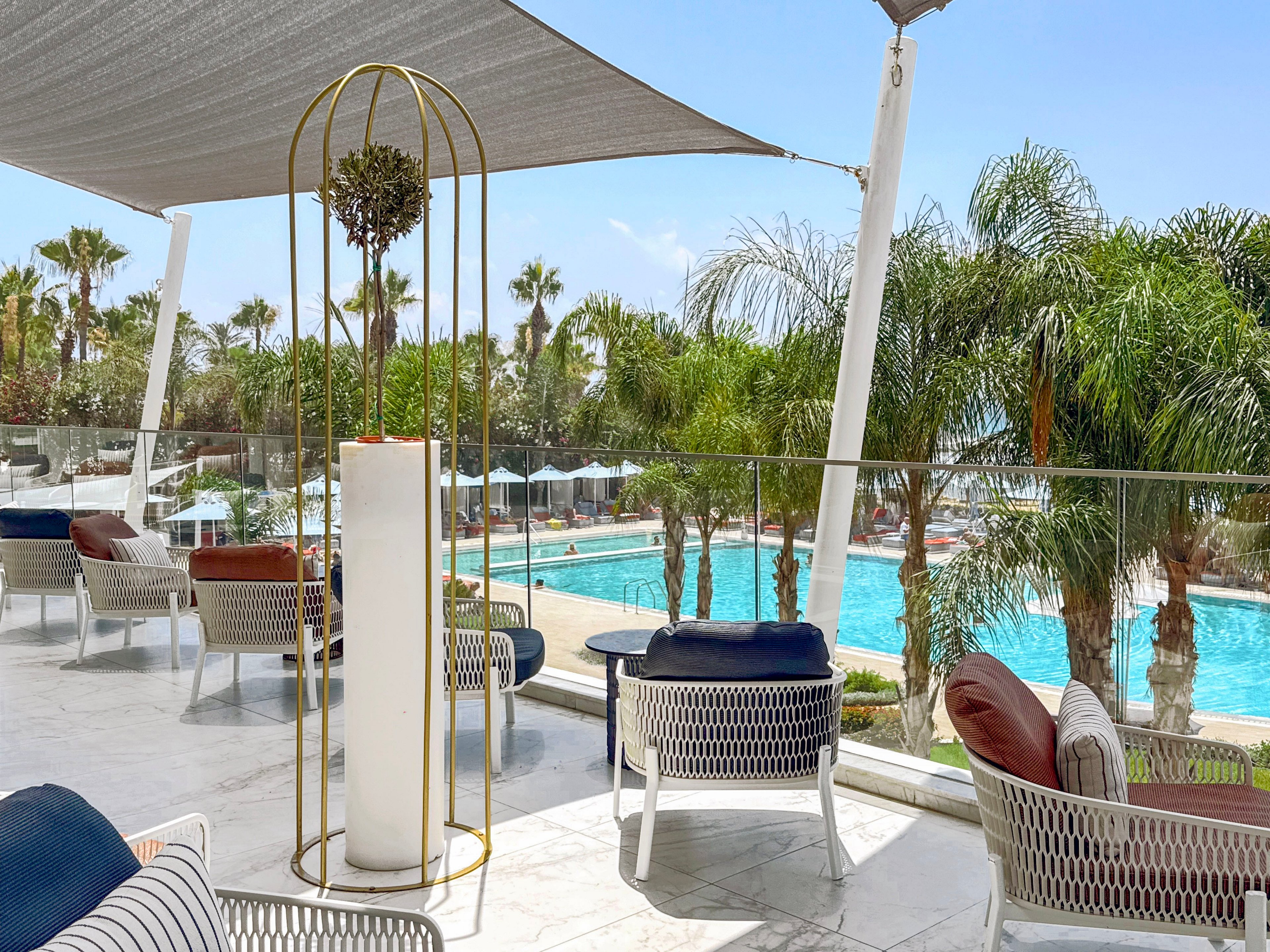 An elegant outdoor lounge terrace overlooks a large turquoise swimming pool surrounded by palm trees. White woven chairs with colorful cushions are arranged on a marble floor beneath a modern shade canopy. A tall decorative planter with a metal frame stands in the foreground, adding a stylish focal point. Sun loungers and umbrellas line the pool area, suggesting a relaxing resort or hotel setting under clear blue skies.