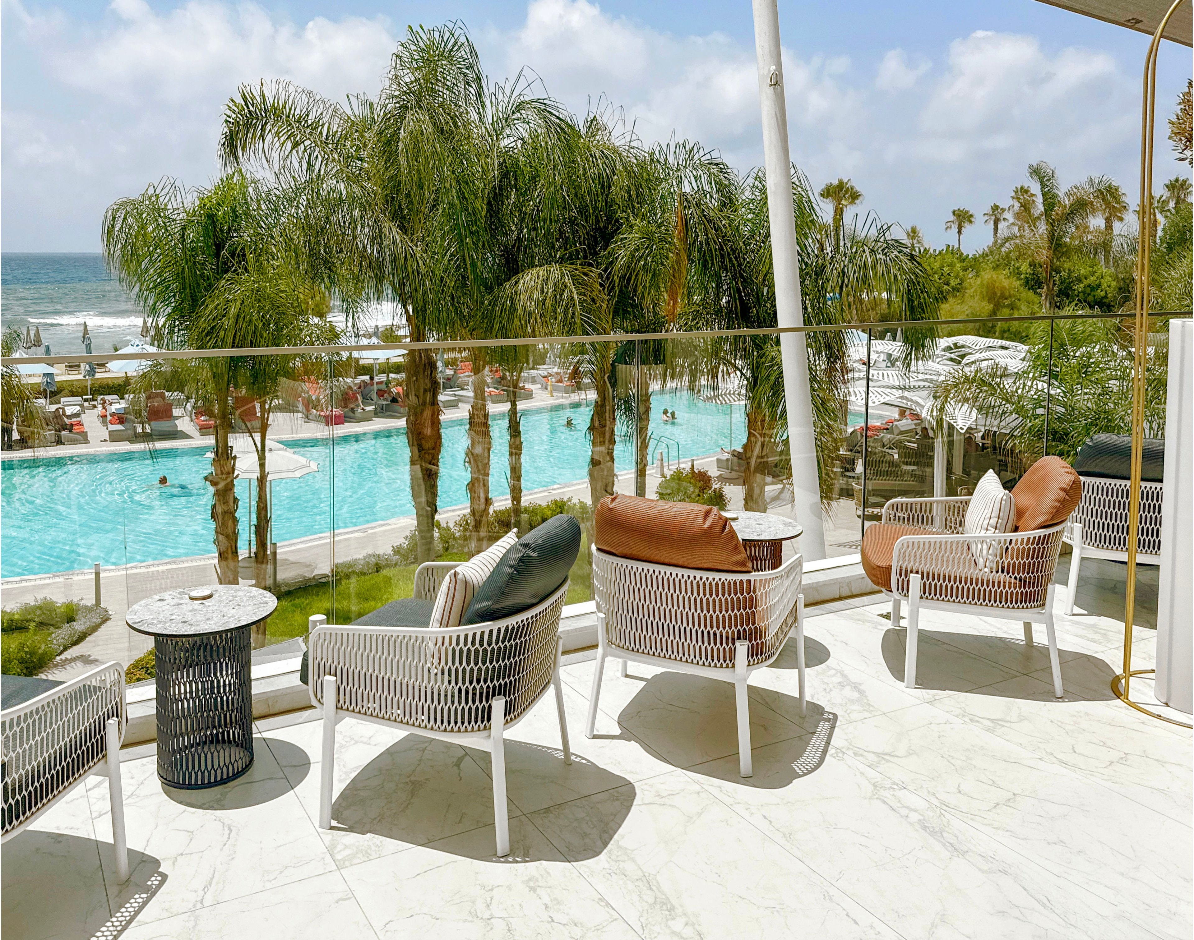 A modern terrace with woven lounge chairs overlooks a turquoise resort swimming pool and the ocean beyond. Palm trees line the pool area, creating a lush tropical atmosphere under a bright, partly cloudy sky. Cushioned chairs and side tables on the marble floor suggest a comfortable space for relaxation and leisure. People can be seen enjoying the pool and sun loungers in the background, enhancing the vacation setting.