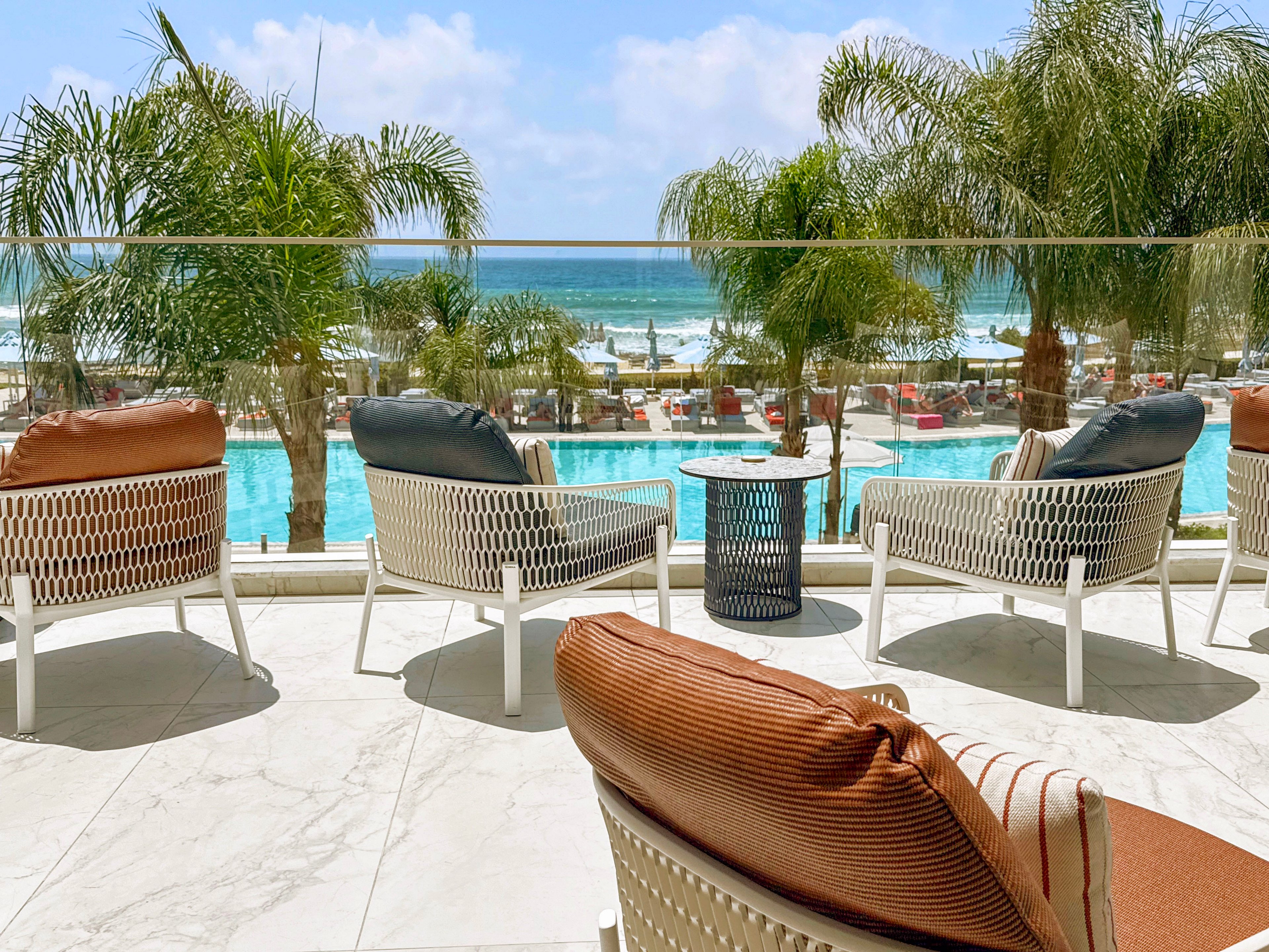 Outdoor lounge chairs sit on a sunlit terrace facing a bright blue swimming pool and the ocean beyond. Palm trees line the pool area, adding a tropical resort atmosphere to the scene. Cushioned wicker chairs and a small side table create a relaxed seating arrangement for guests. The clear sky and calm water emphasize a warm, leisurely vacation mood.