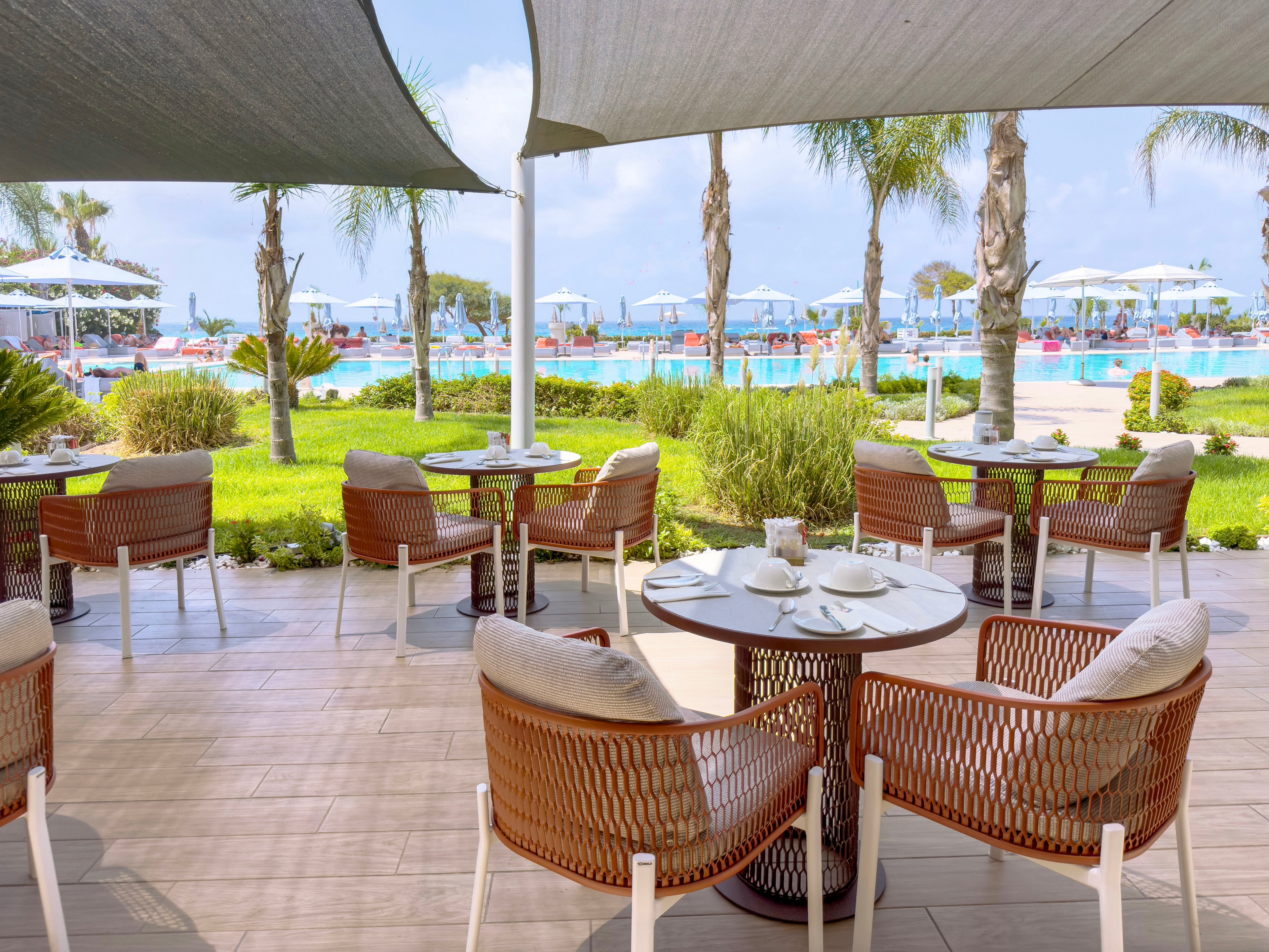 An elegant outdoor café terrace overlooks a large swimming pool at a resort. Wicker chairs and round tables are neatly arranged under shade sails, set with white plates and cups. Palm trees, green lawn, and sun loungers with umbrellas frame the bright blue water. The scene suggests a relaxed vacation atmosphere on a sunny day.