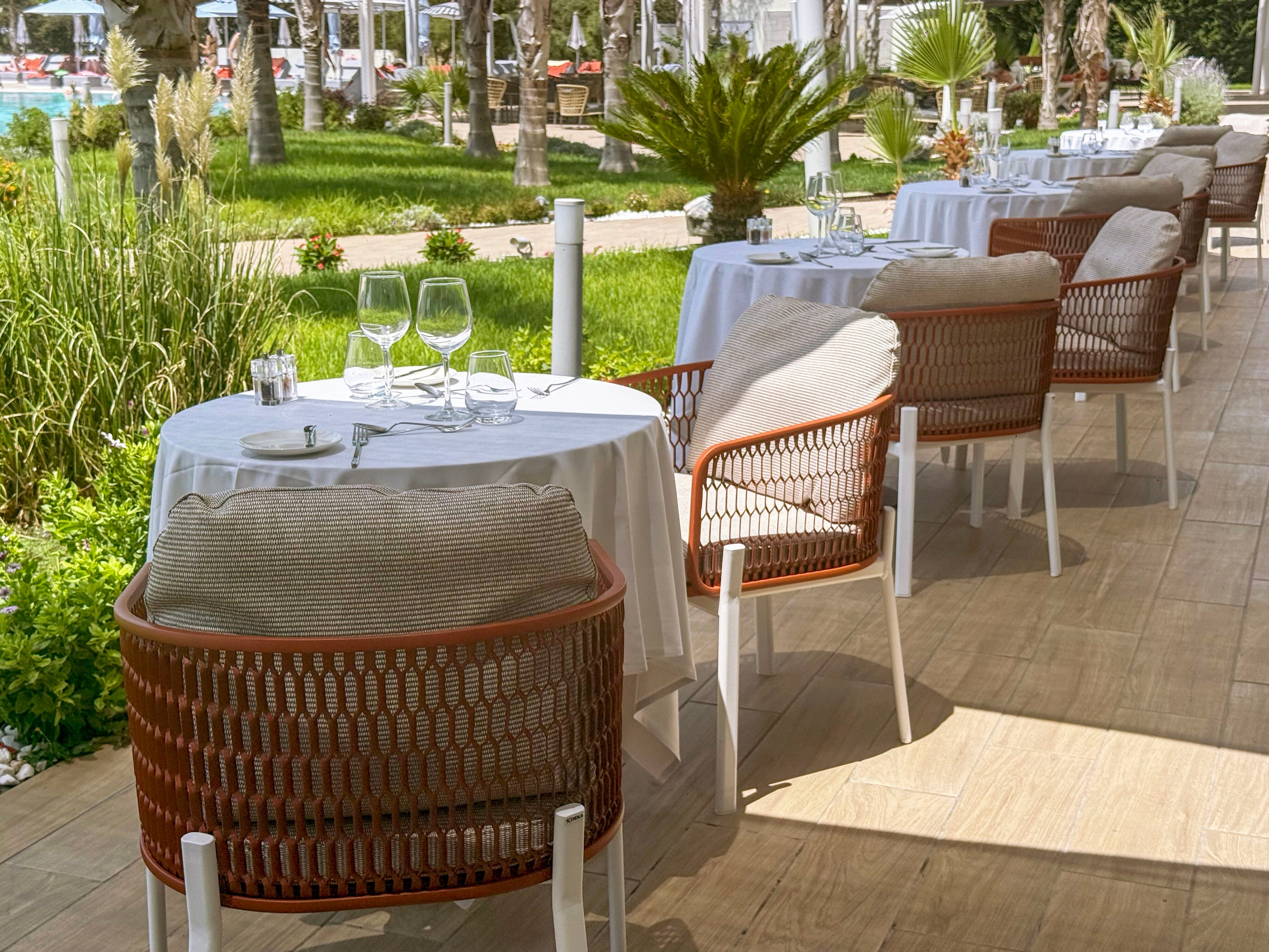 An inviting outdoor restaurant terrace features neatly arranged tables with white tablecloths and cushioned wicker chairs. Each table is set with clear wine glasses, water glasses, and small items, ready for guests. The terrace overlooks a lush green garden with palm trees and manicured plants, creating a relaxed vacation atmosphere. Sunlit wooden flooring and soft neutral tones contribute to a calm, upscale dining experience.