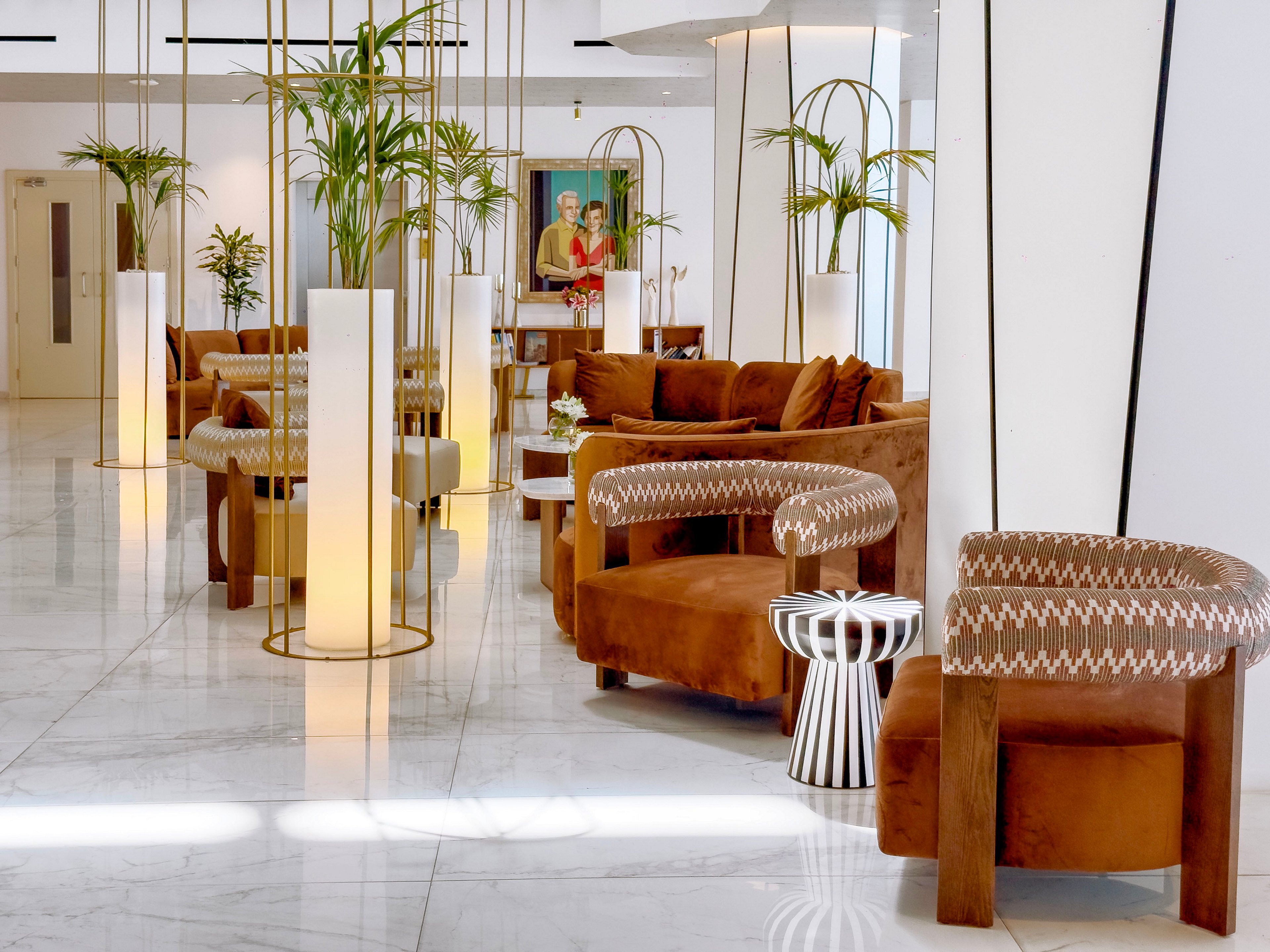A contemporary lobby features brown velvet armchairs and sofas arranged on a glossy white marble floor. Tall cylindrical planters with green plants and warm lighting create a welcoming atmosphere. Decorative side tables and patterned chair backs add visual interest to the modern interior design. In the background, a framed portrait painting hangs on the wall, enhancing the sophisticated setting.