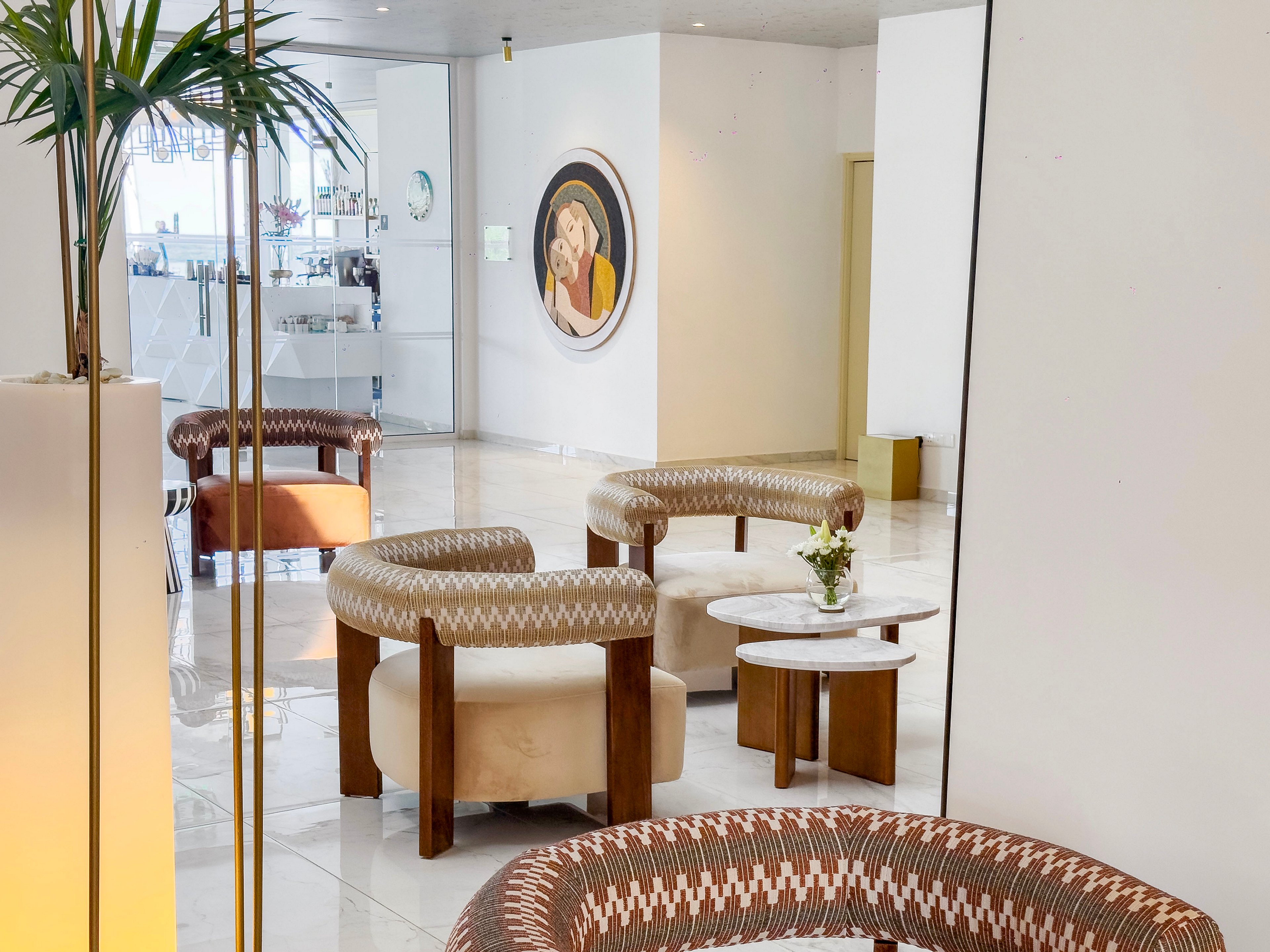 A contemporary lounge area features circular armchairs with geometric patterned upholstery and wooden legs. The space has a bright, minimalist interior with glossy white floors and light-colored walls. A small round marble table with a flower arrangement sits between the chairs, adding a decorative touch. In the background, a framed artwork and potted plants contribute to the stylish, relaxed atmosphere.