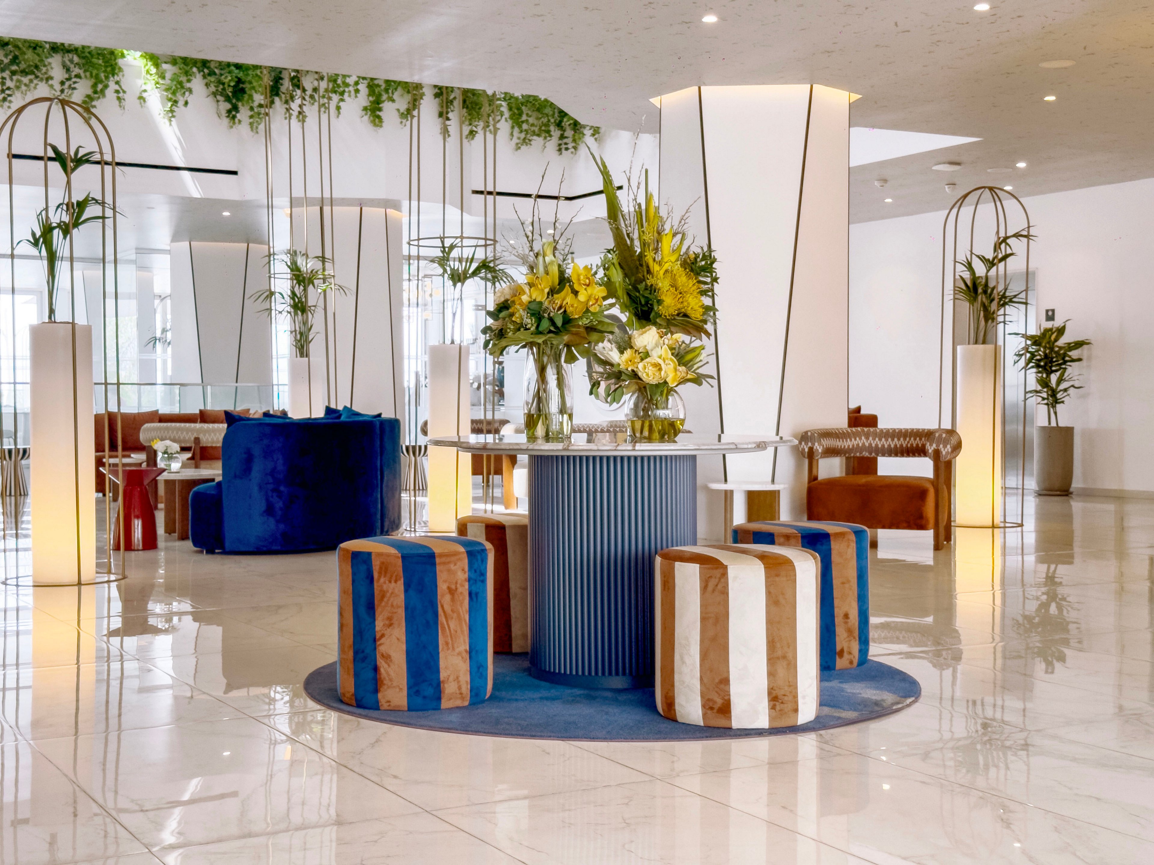 A contemporary lobby features a round blue rug with striped ottomans surrounding a central table topped with fresh floral arrangements. The space is bright and airy, with glossy marble flooring and tall white light columns adding vertical emphasis. Curved sofas and decorative metal arches frame the seating area, while greenery hangs from the ceiling and potted plants soften the modern design. The overall atmosphere is stylish, welcoming, and sophisticated.