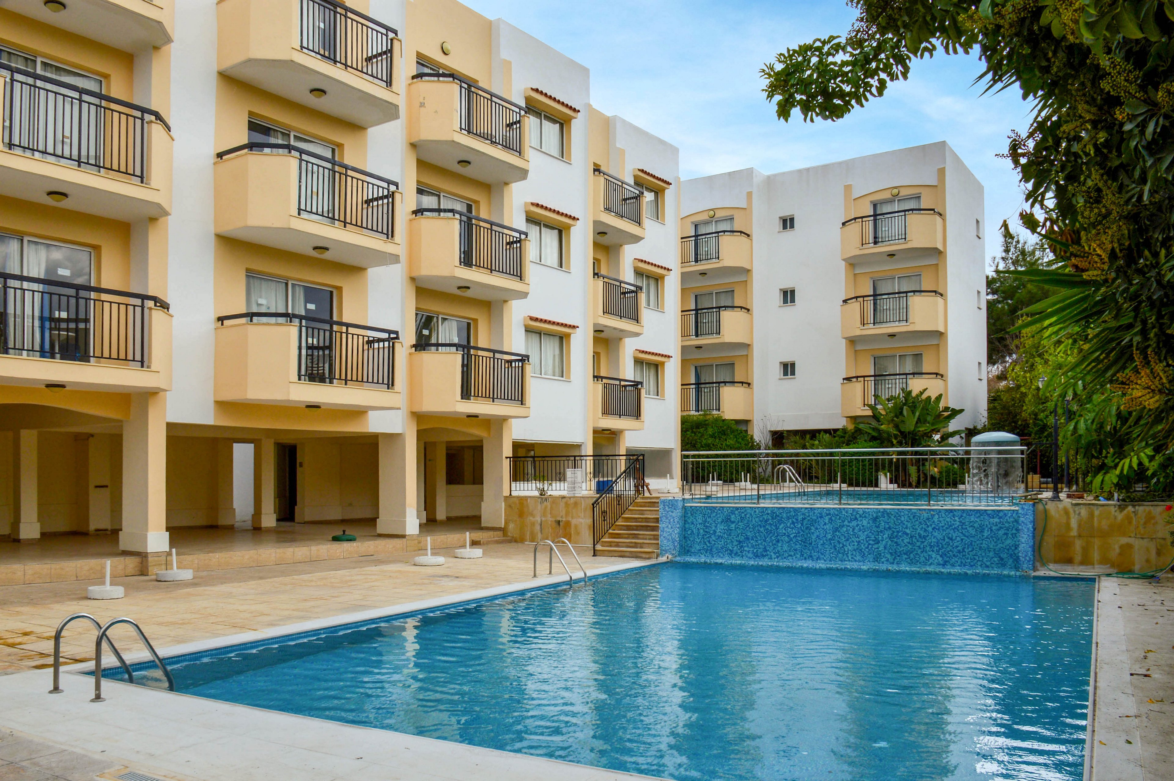 The image shows several contemporary apartment buildings with balconies, situated around a spacious outdoor swimming pool. The pool area features tiled flooring and metal ladders, with greenery visible in the background. The scene is calm and overcast, highlighting the clean architectural lines and inviting communal space.