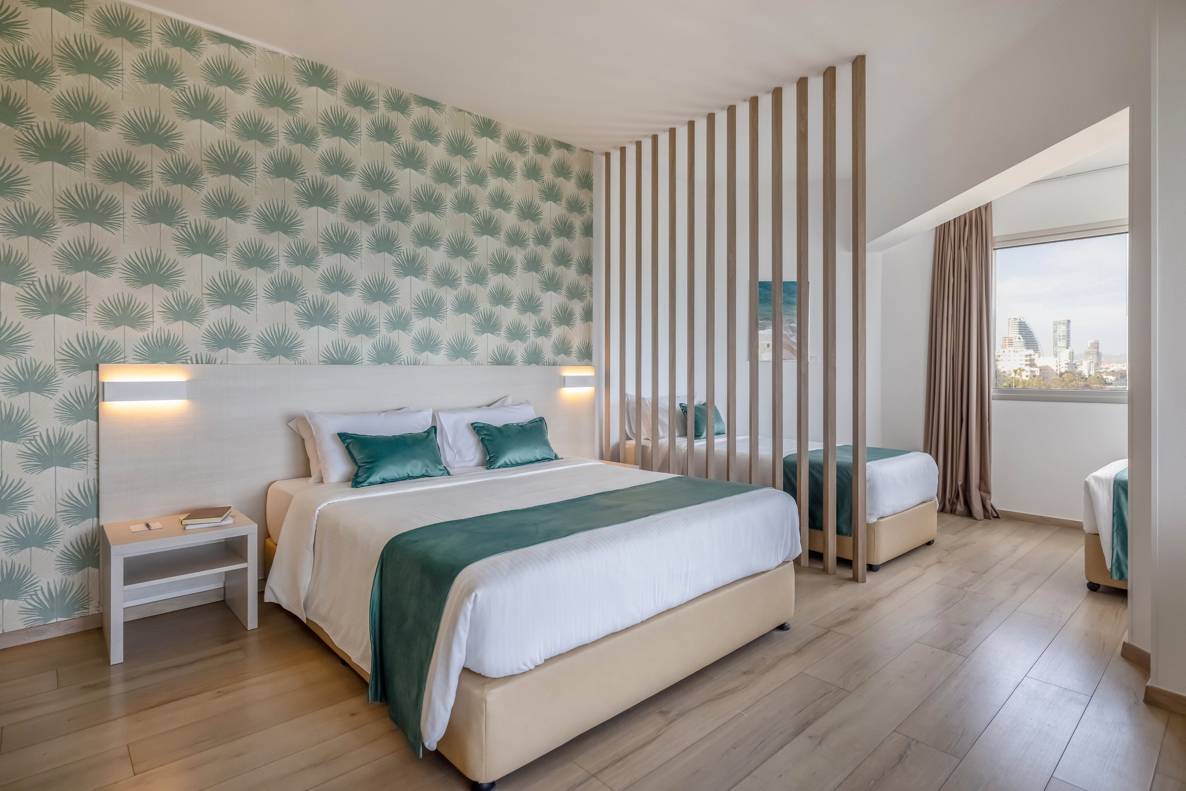 A contemporary hotel bedroom features a large double bed with teal accents and matching pillows. The space is divided by vertical wooden slats, separating the main bed from an additional single bed. Light wood flooring and a patterned accent wall with leaf motifs create a fresh, coastal-inspired look. A window at the back reveals a distant city skyline under a bright sky.