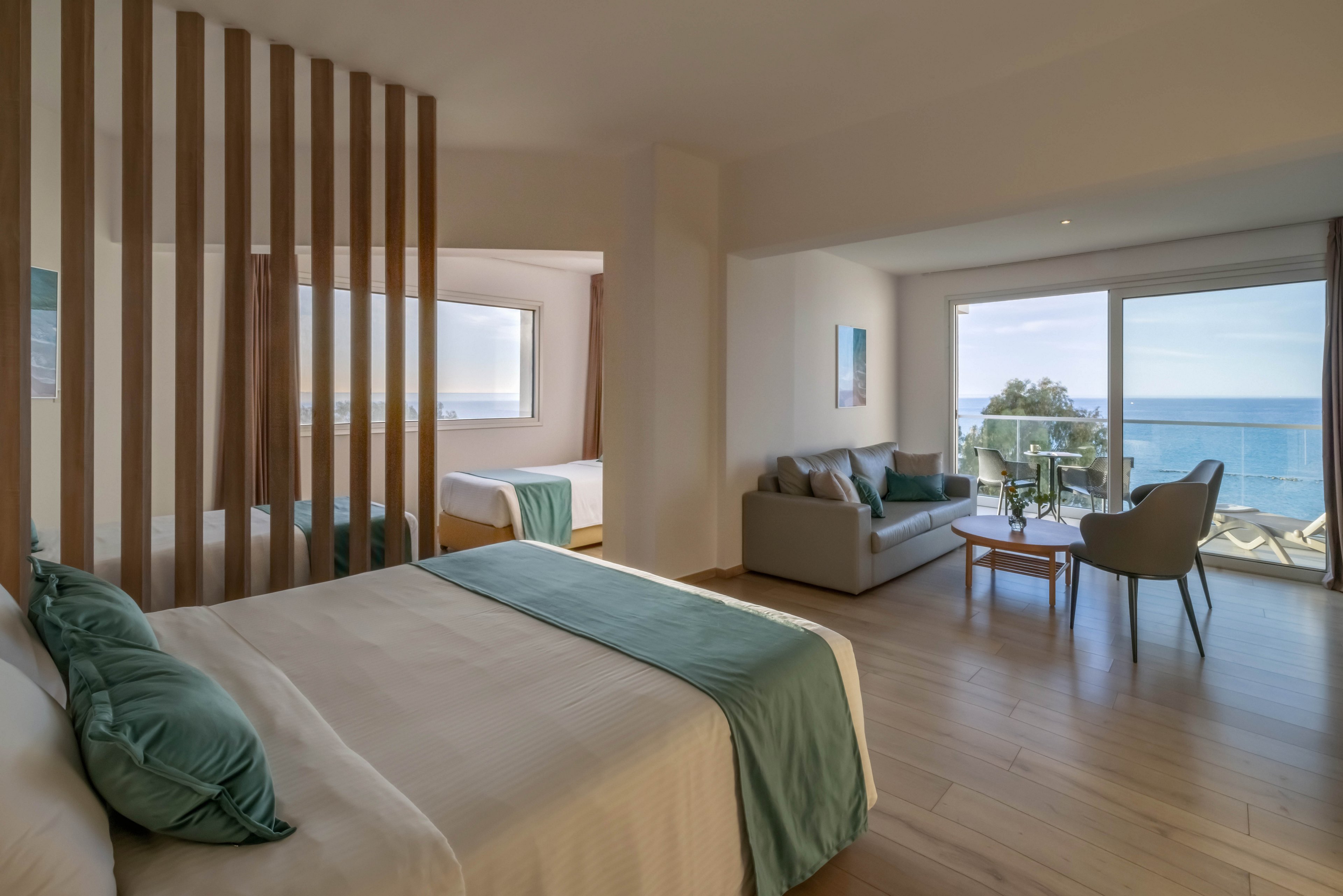 A contemporary hotel suite features a large bed in the foreground with soft white linens and teal accents. A wooden slatted partition separates the main sleeping area from an additional bed near a window. The room opens onto a balcony through large glass doors, revealing a clear view of the sea and coastline. A cozy seating area with a sofa, chairs, and small table completes the relaxed, coastal atmosphere.