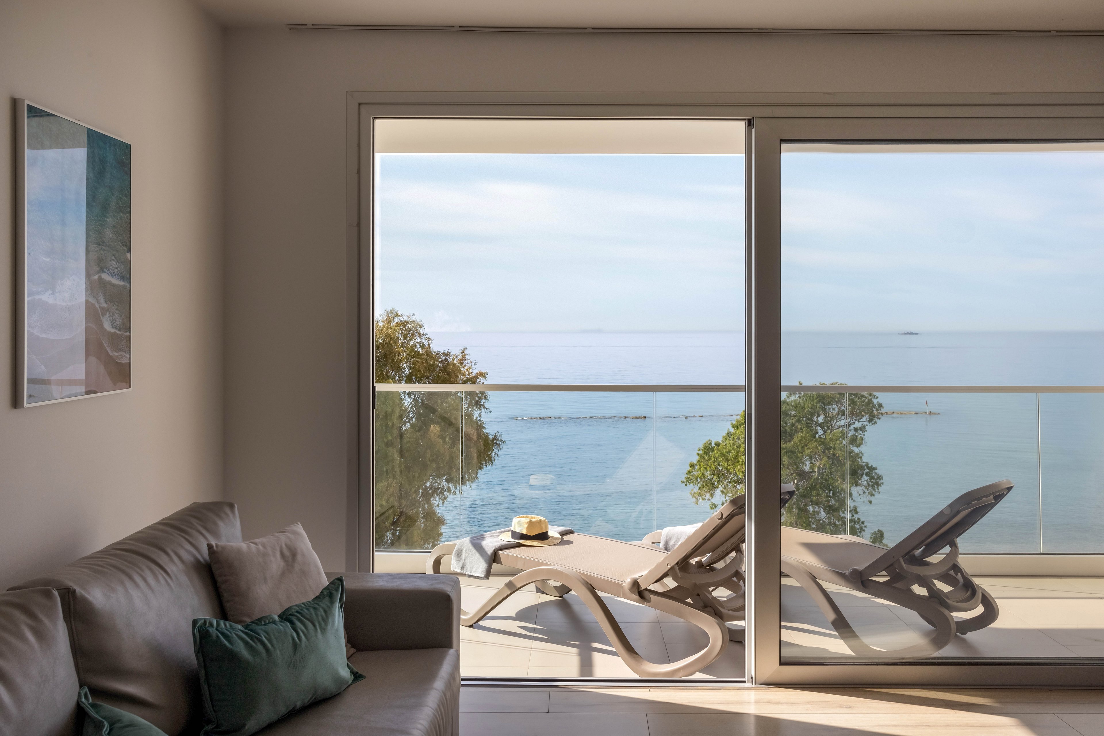 A contemporary apartment living room opens onto a balcony overlooking the sea. Two sun loungers sit on the terrace beside a small table holding a hat and drink. Large sliding glass doors frame the coastal view with calm blue water and distant horizon. Soft neutral tones and minimal decor create a relaxed, vacation-like atmosphere.