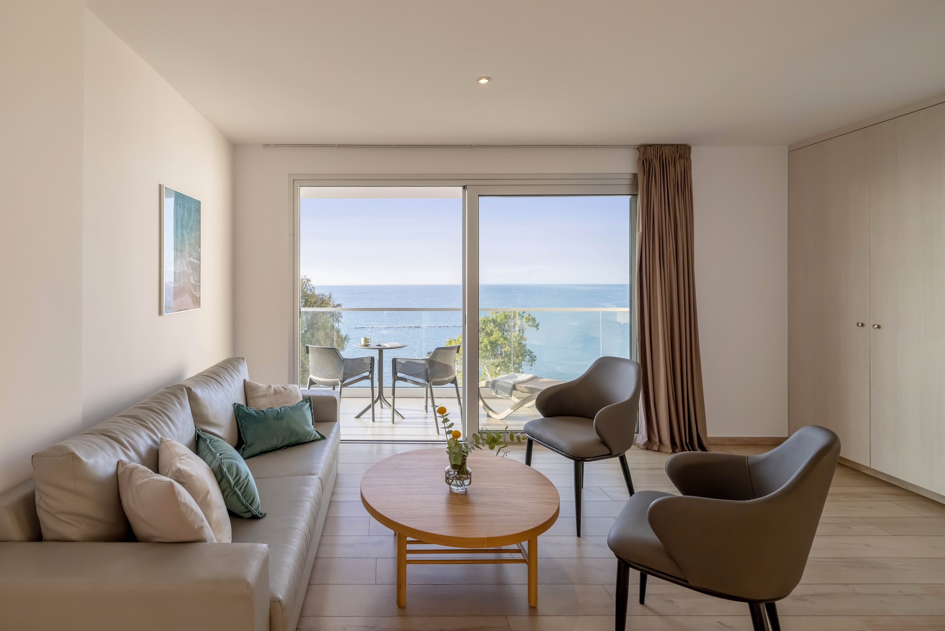 A contemporary living room features a neutral-toned sofa and two armchairs arranged around a round wooden coffee table. Large sliding glass doors open to a balcony with a small table and chairs overlooking the ocean. Soft cushions, a simple wall art piece, and a vase with flowers add subtle decorative touches. Natural light fills the space, creating a calm and inviting coastal atmosphere.