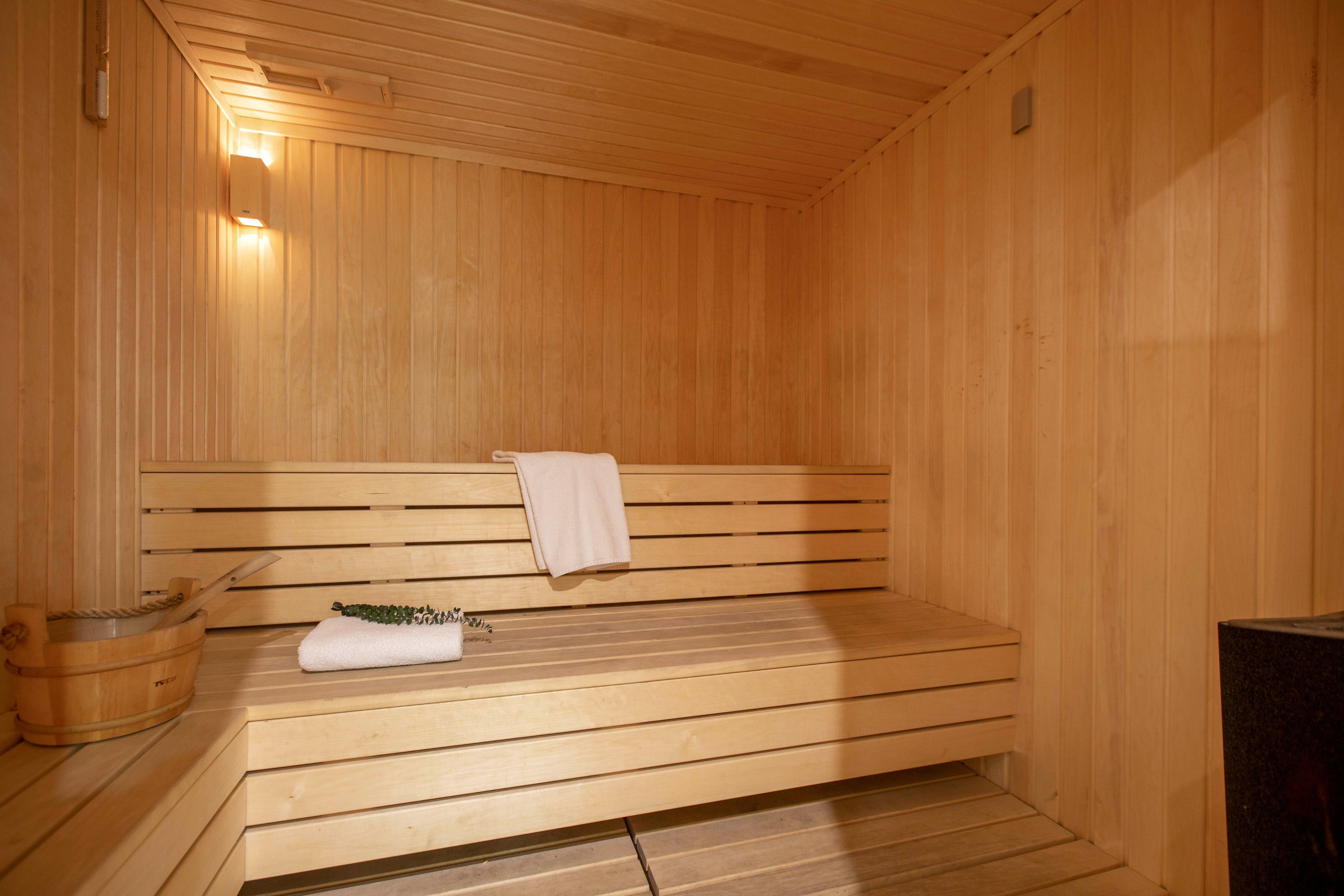 A warm wooden sauna interior features a tiered bench and soft ambient lighting. A white towel is draped over the upper bench, suggesting recent or upcoming use. A wooden bucket and ladle sit on the floor near the corner, ready for creating steam. The light wood panels and simple design create a clean, minimalist spa atmosphere.