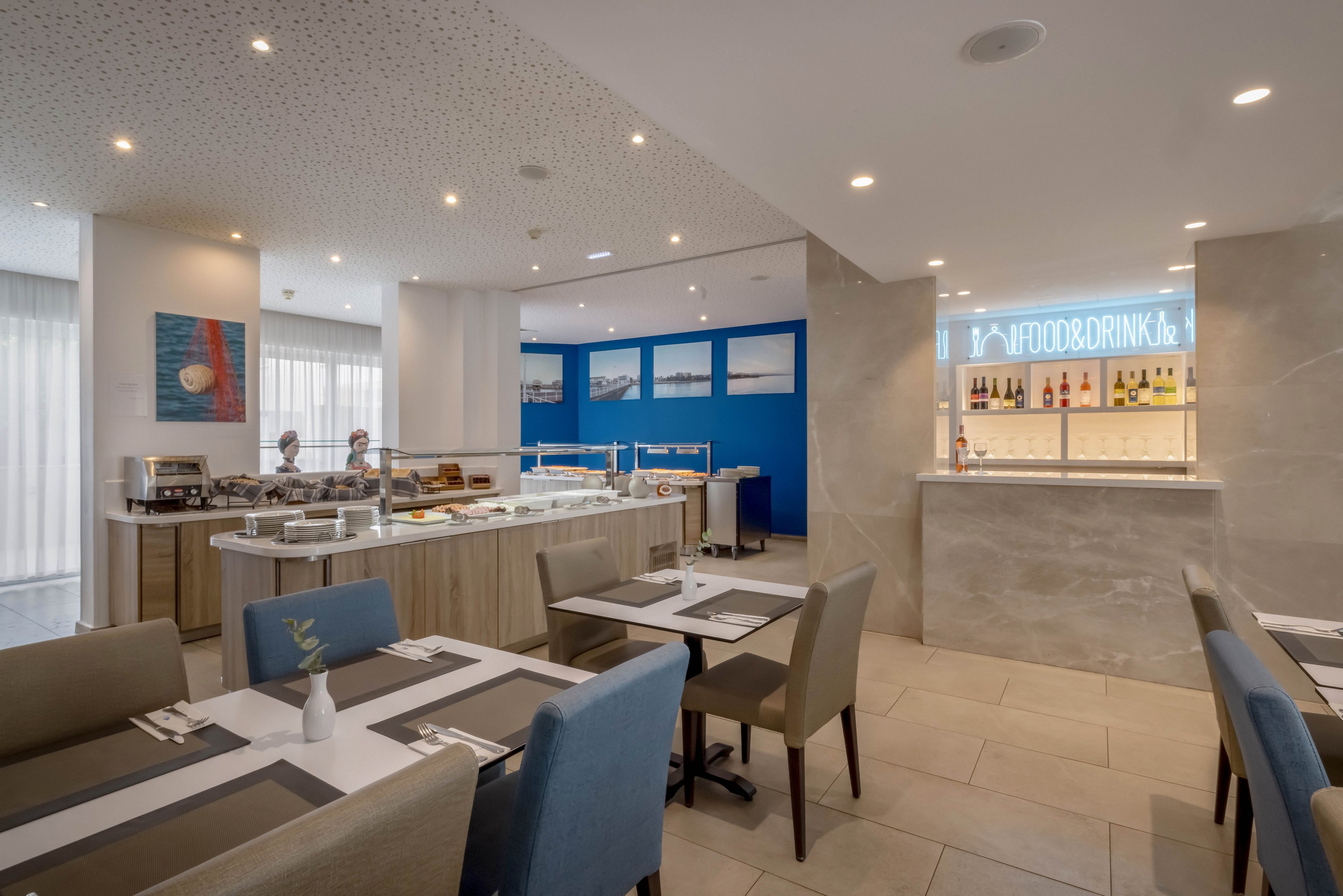 Contemporary dining room featuring a self-service buffet counter in a hotel or restaurant. Neatly arranged tables with placemats and cutlery face the buffet, which displays various covered food items. A bright blue accent wall and framed artwork add color to the neutral-toned interior. In the background, a bar area with illuminated signage and bottles provides additional food and drink options. Recessed ceiling lights and large windows create a clean, well-lit atmosphere.