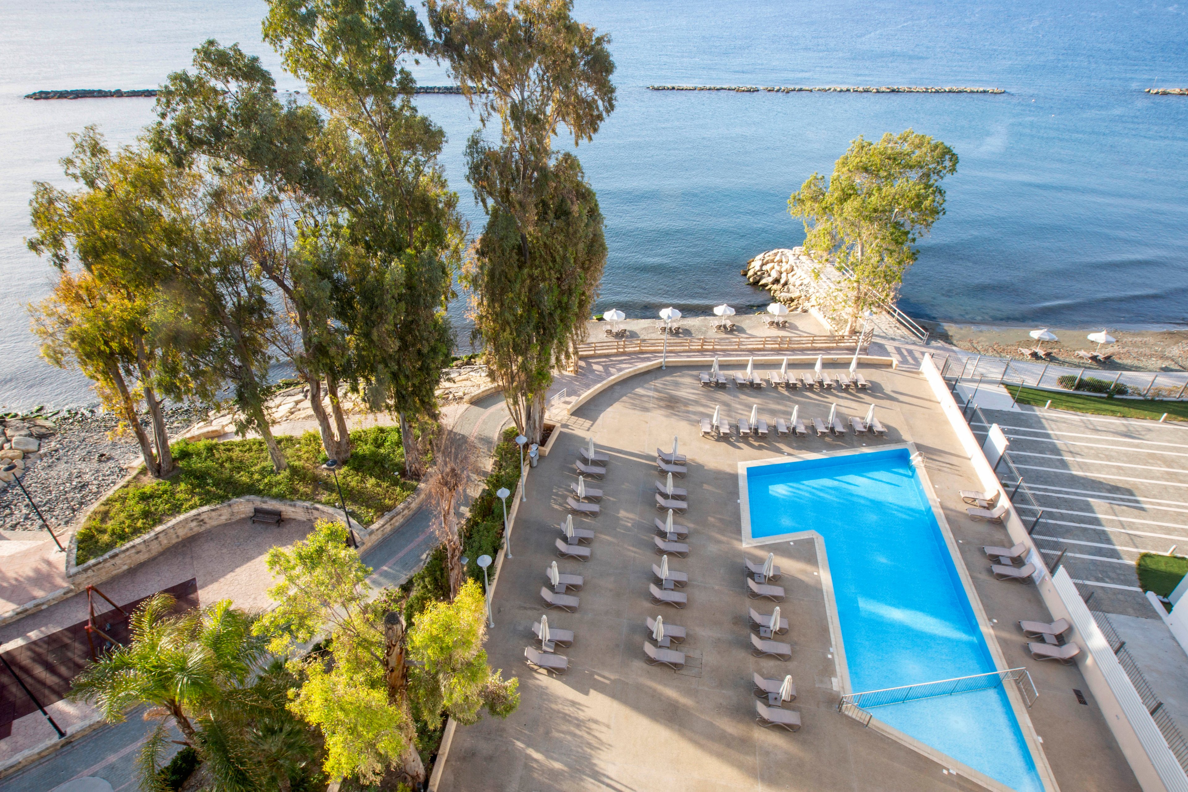 An elevated view shows a hotel pool terrace beside a calm seaside shoreline. Numerous sun loungers are neatly arranged around a bright blue, angular swimming pool. Tall trees provide some natural shade, while the sea and a rocky breakwater stretch into the distance. The scene is quiet and orderly, suggesting a peaceful resort environment ready for guests.