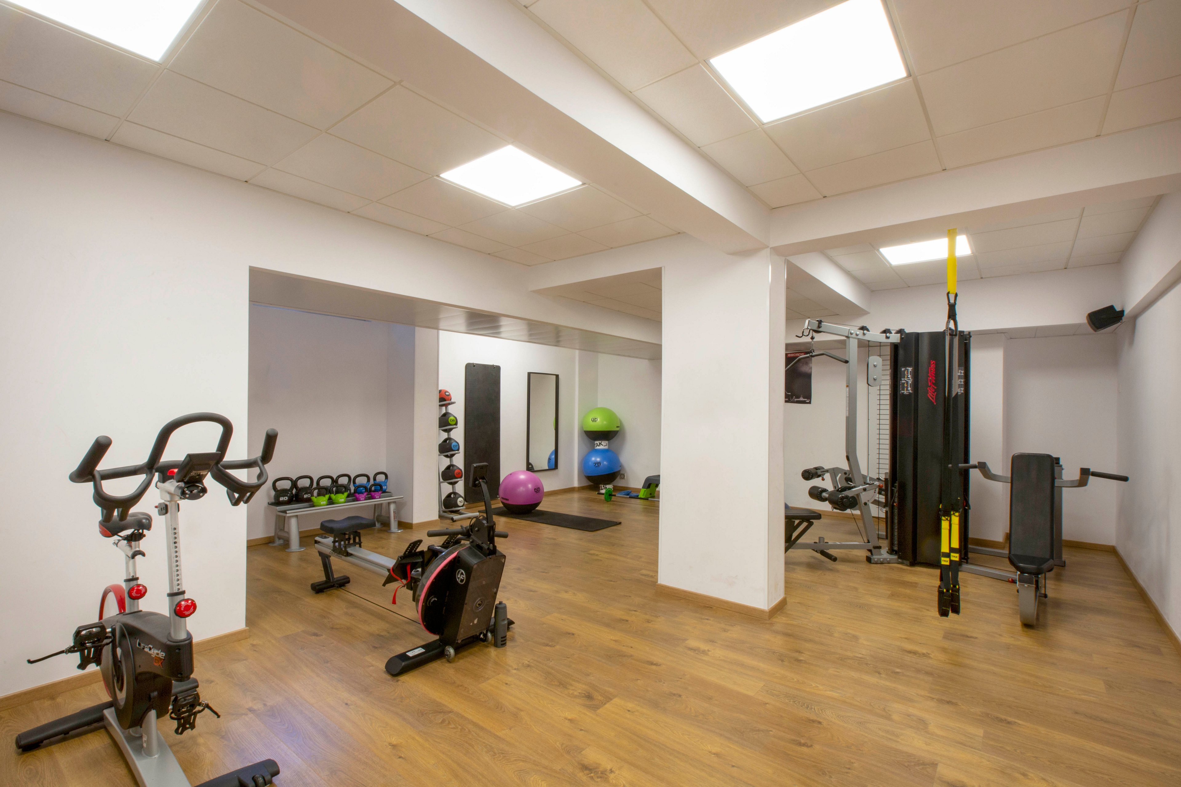 A clean, modern indoor gym features various exercise machines and free weights arranged neatly on a wooden floor. Stationary bikes, a weight machine, and suspension straps are visible in the foreground. In the background, colorful kettlebells, stability balls, and benches line the walls. Bright ceiling lights and white walls create a spacious, well-lit workout environment.
