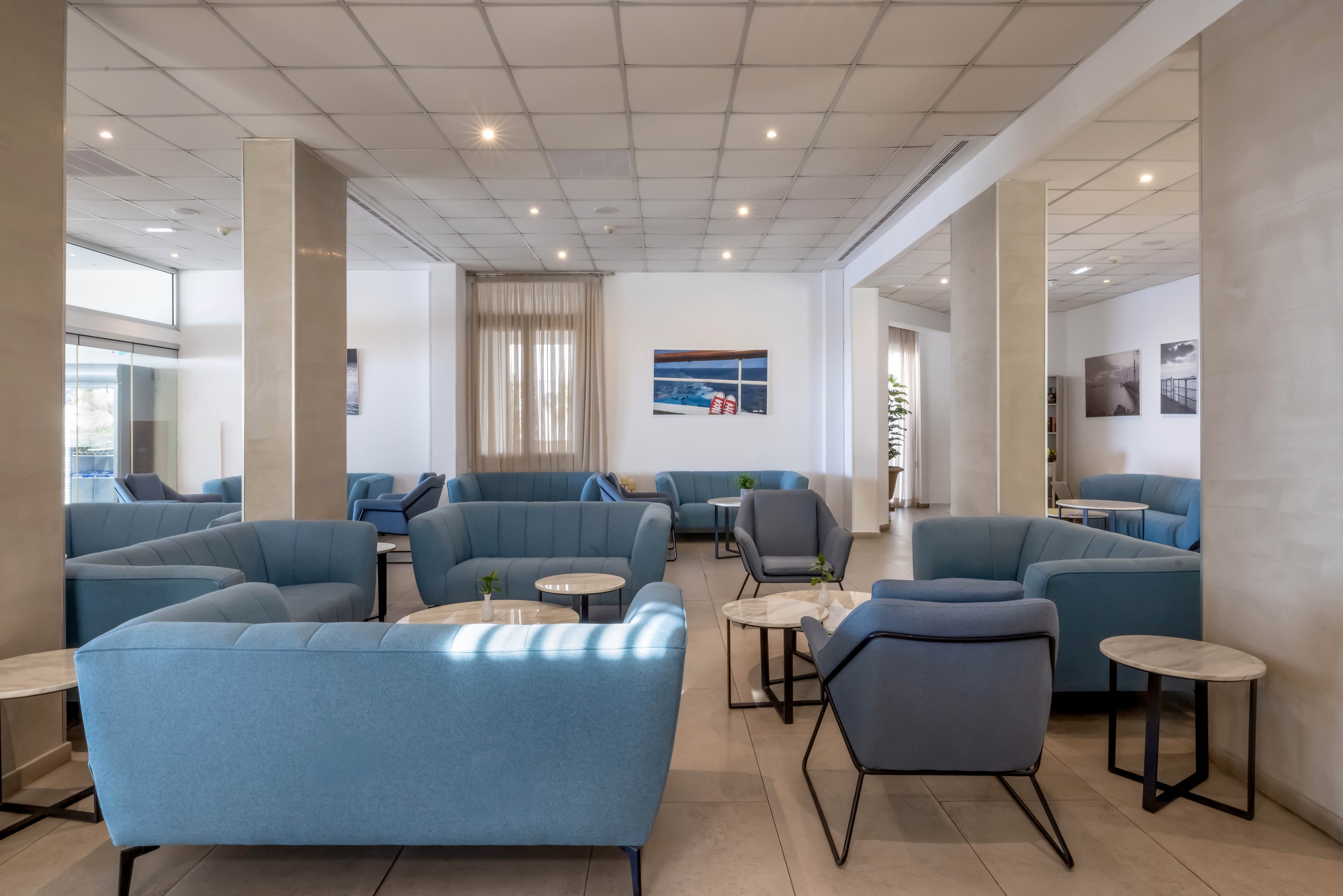 A spacious modern lounge features multiple light blue sofas and armchairs arranged around small round tables. The room has a neutral color palette with beige floors, white walls, and a grid-patterned ceiling with recessed lighting. Large windows and glass doors allow natural light to brighten the seating area. Framed artwork decorates the walls, adding subtle visual interest to the contemporary interior.