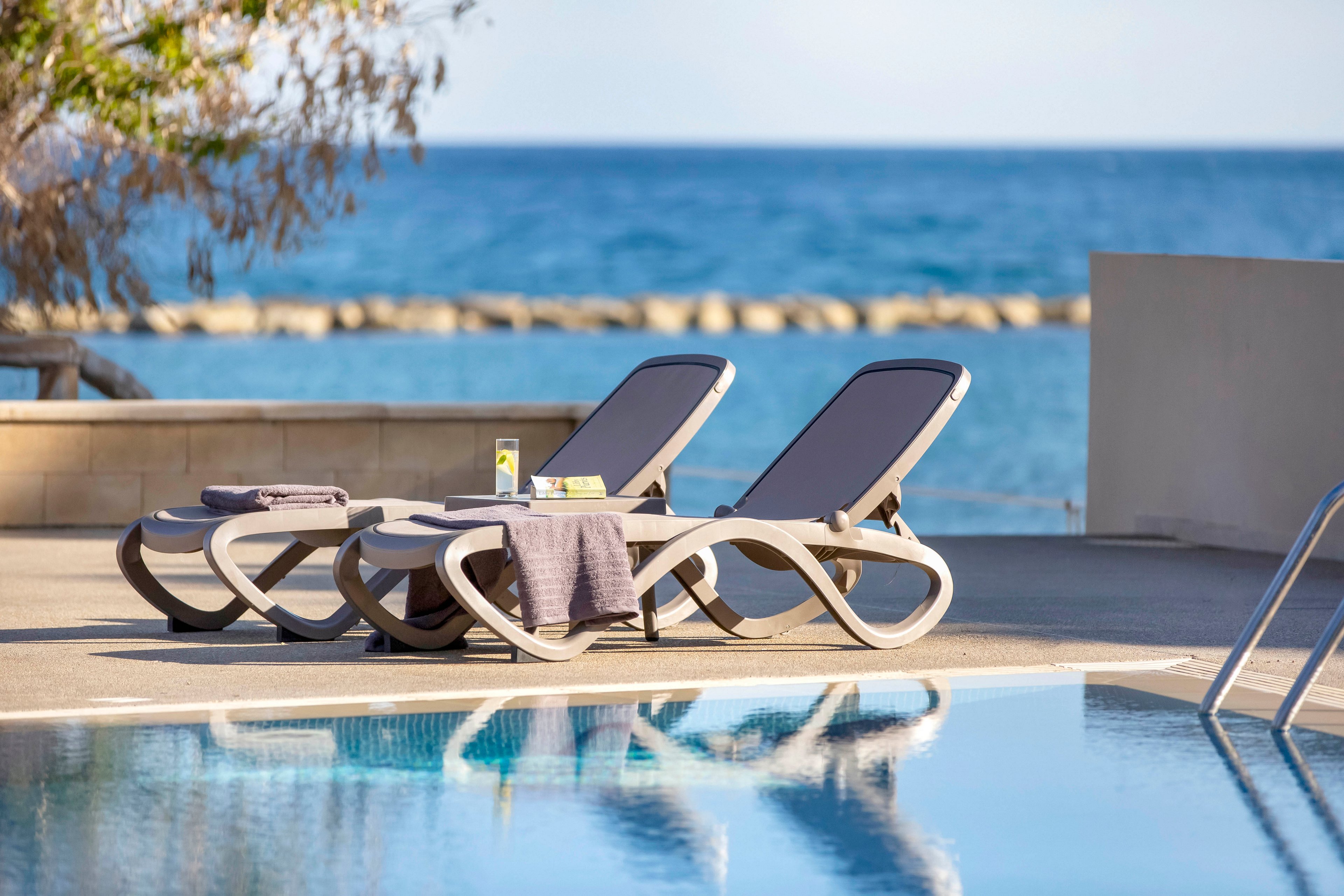 Two contemporary sun loungers are arranged beside a calm swimming pool with a clear reflection of the chairs in the water. A small side table between the loungers holds a drink and other small items, suggesting a relaxing break. A folded towel is draped over one lounger, while another towel rests on the table. In the background, the blue ocean and a low stone breakwater create a serene coastal setting under clear skies.
