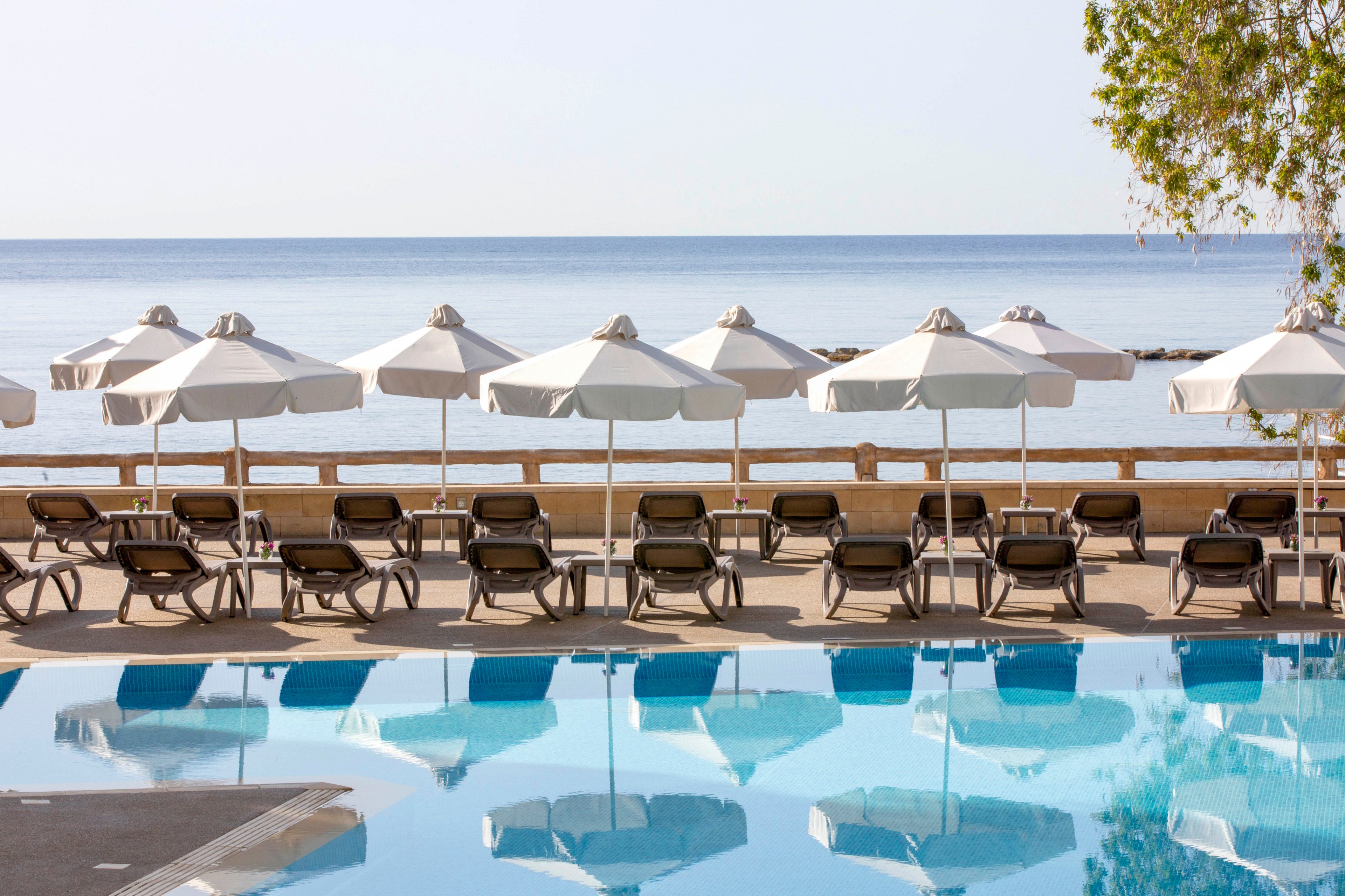 A tranquil resort pool sits in the foreground, reflecting rows of white sun umbrellas and lounge chairs. Beyond the pool, a wooden railing separates the deck area from the calm, blue sea. The scene is bright and sunny, with clear skies and gentle light suggesting a peaceful vacation setting. A tree on the right edge adds a touch of greenery to the otherwise minimalist coastal view.