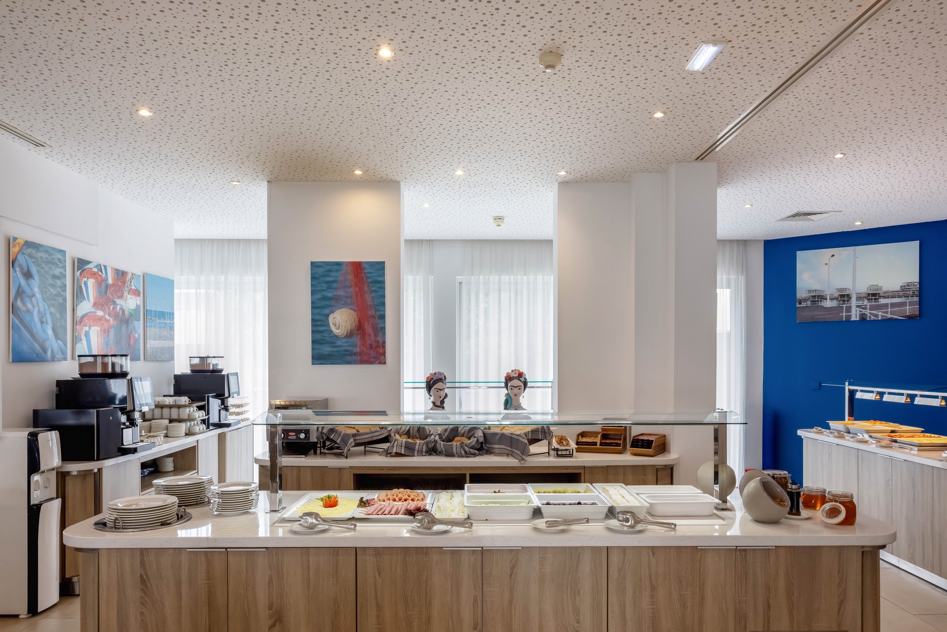 A contemporary breakfast buffet is set up in a bright hotel dining area. The foreground counter displays plates, bowls, and trays of cold cuts, cheese, and fresh fruit. Behind it, a second counter holds pastries, bread, and cereal dispensers, with coffee machines on the left. Large windows with sheer curtains and framed artwork decorate the walls, creating a clean and inviting atmosphere. The overall scene feels organized, modern, and ready for guests.