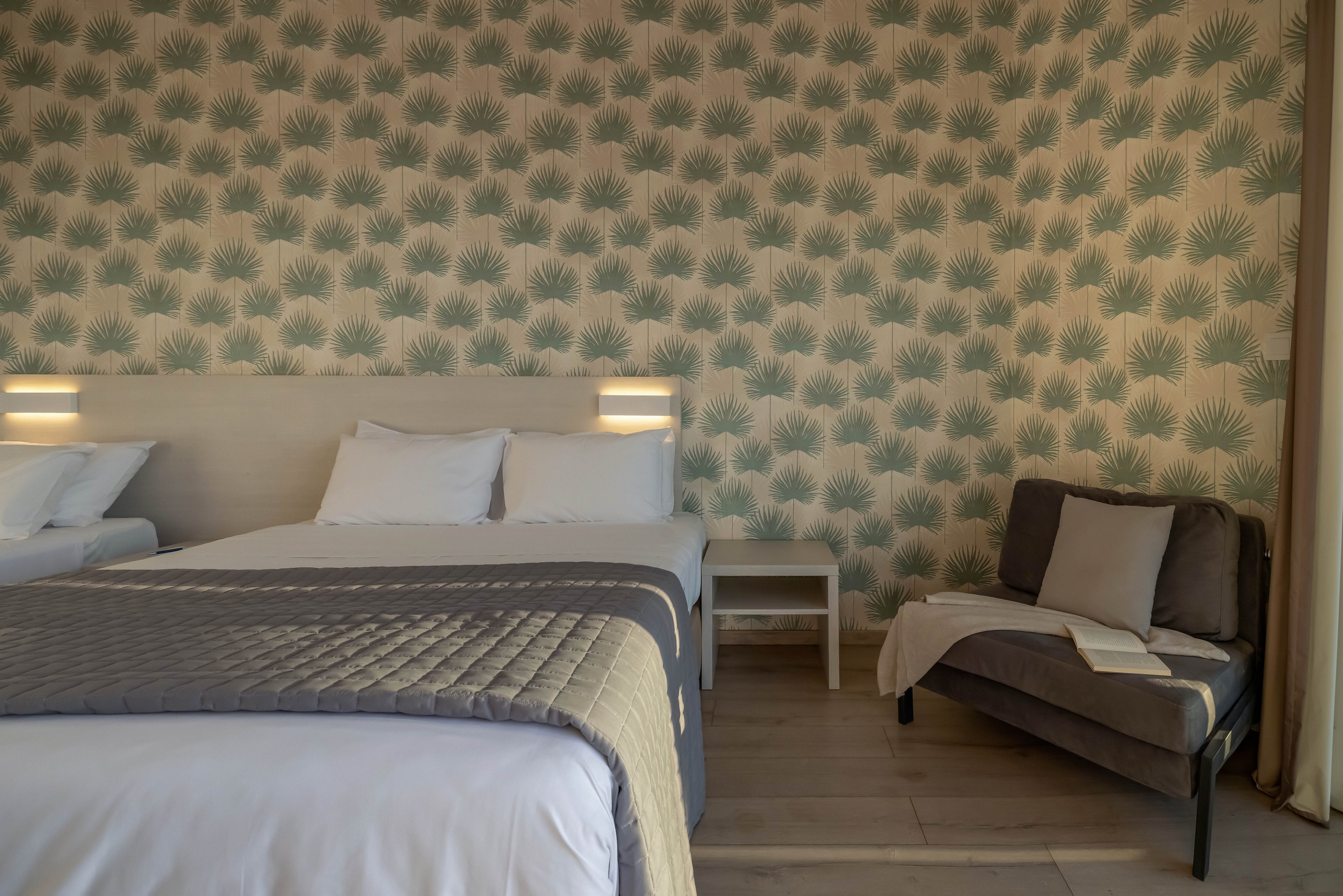 A neatly made double bed with gray quilted bedding stands against a wall covered in a subtle leaf-patterned wallpaper. Soft wall-mounted lights create a warm and inviting atmosphere in the room. To the side, a small sofa with cushions and a draped blanket offers an additional seating or lounging area. A simple bedside table and light wooden flooring complete the clean, contemporary interior design.
