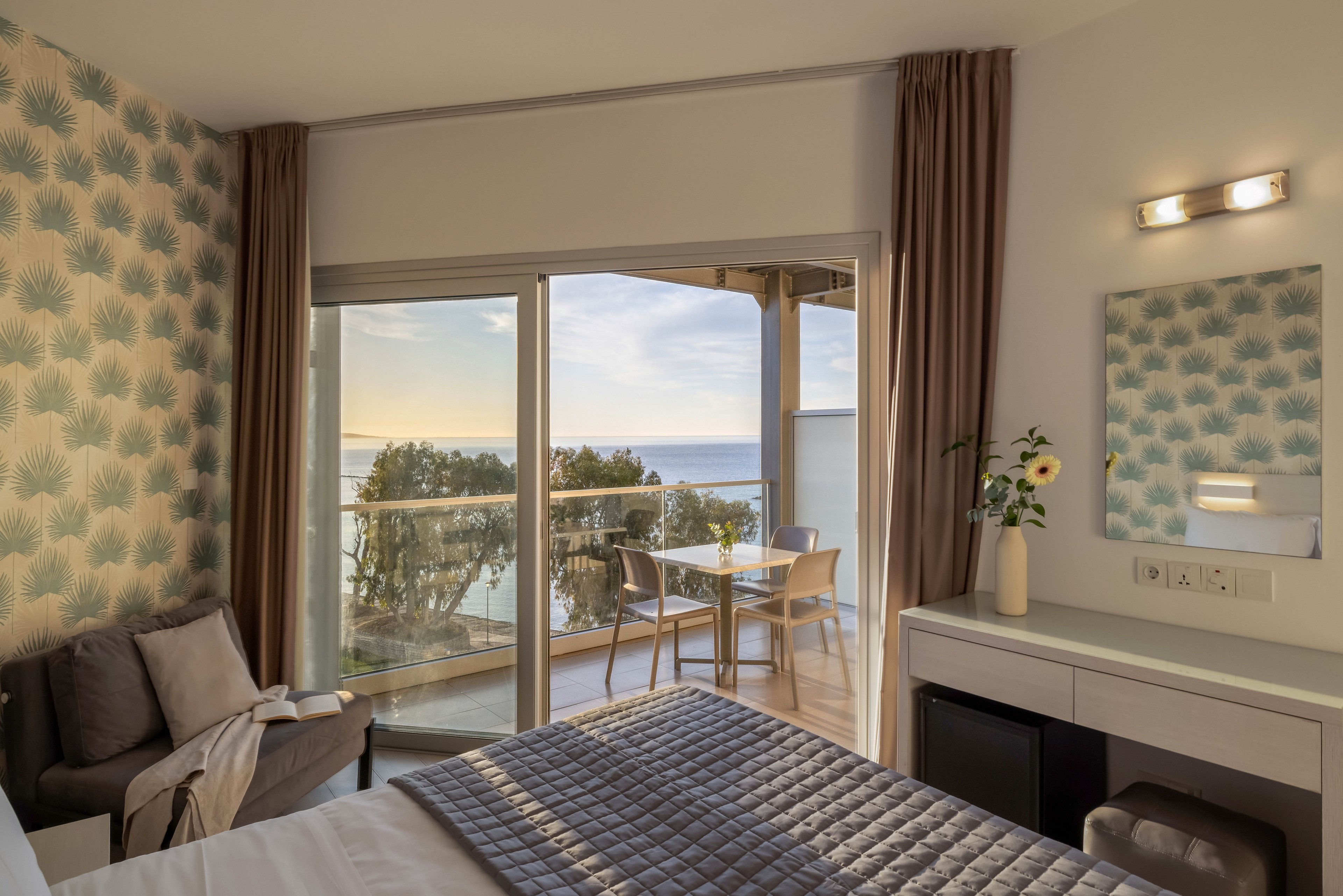 A contemporary hotel bedroom features a neatly made bed with a quilted gray cover and soft pillows. Large sliding glass doors open onto a balcony with a small table and two chairs overlooking the sea and trees. Warm evening light fills the room, highlighting patterned wallpaper, beige curtains, and a compact desk with a mirror and vase of flowers. The overall atmosphere is calm, inviting, and ideal for a relaxing coastal stay.
