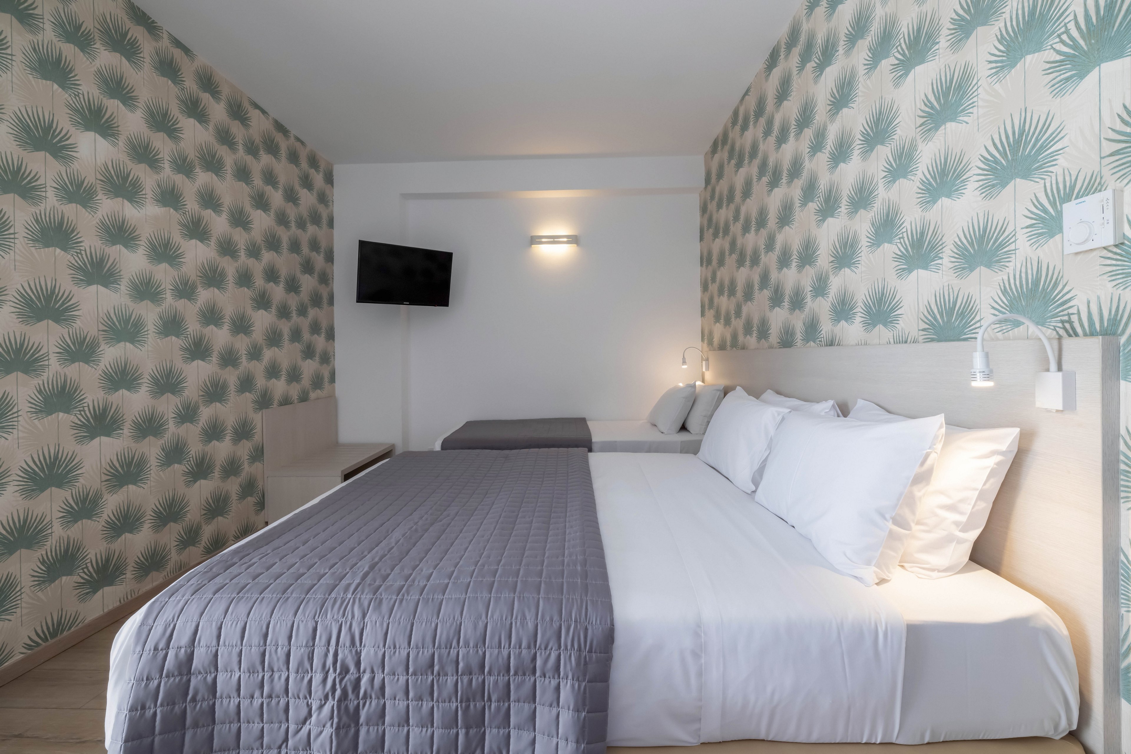 A neatly made double bed with white sheets and a gray quilt dominates a modern hotel room. The space features patterned wallpaper with green palm leaves on both side walls. A wall-mounted flat-screen television and minimalist bedside lamps add contemporary touches. Soft, neutral lighting creates a calm and inviting atmosphere for guests.