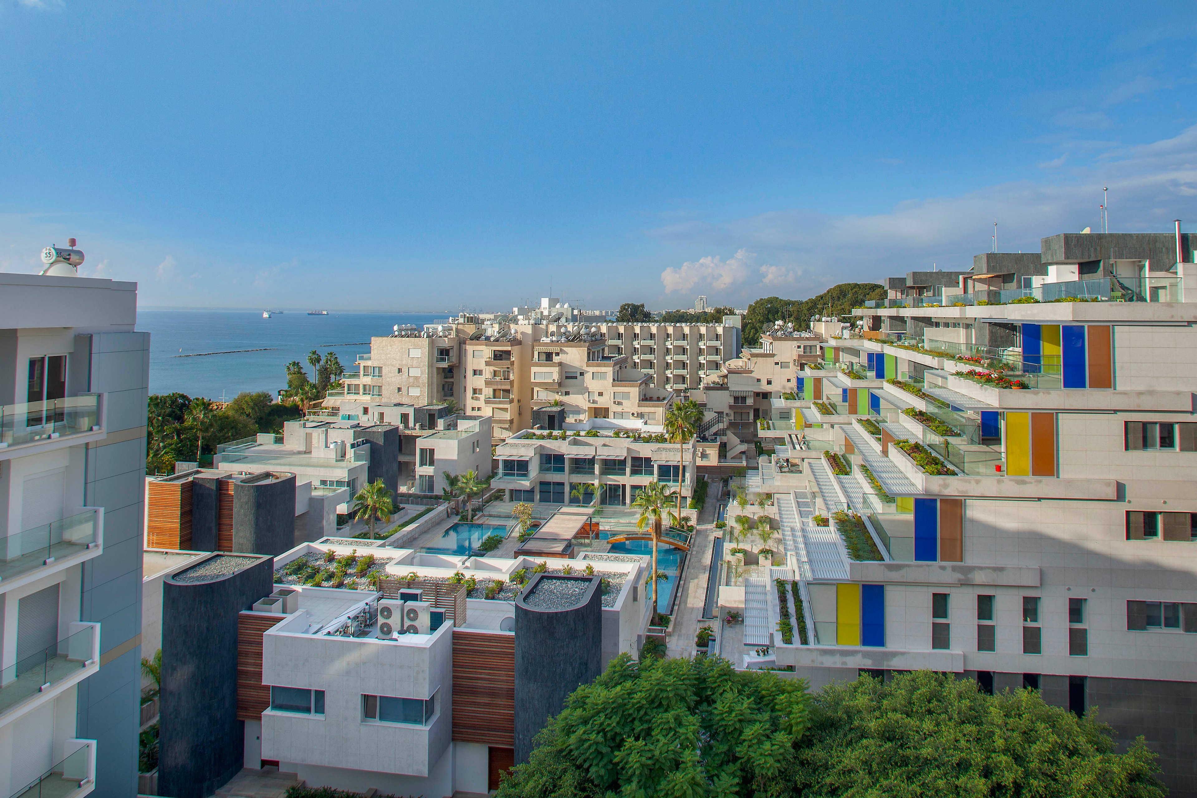 A contemporary apartment complex with multiple white buildings overlooks a calm seaside. The structures feature stepped terraces with greenery and colorful vertical panels in blue, yellow, and red. A central courtyard area includes swimming pools, palm trees, and communal spaces. In the distance, more coastal buildings and the open sea are visible under a clear blue sky with scattered clouds.