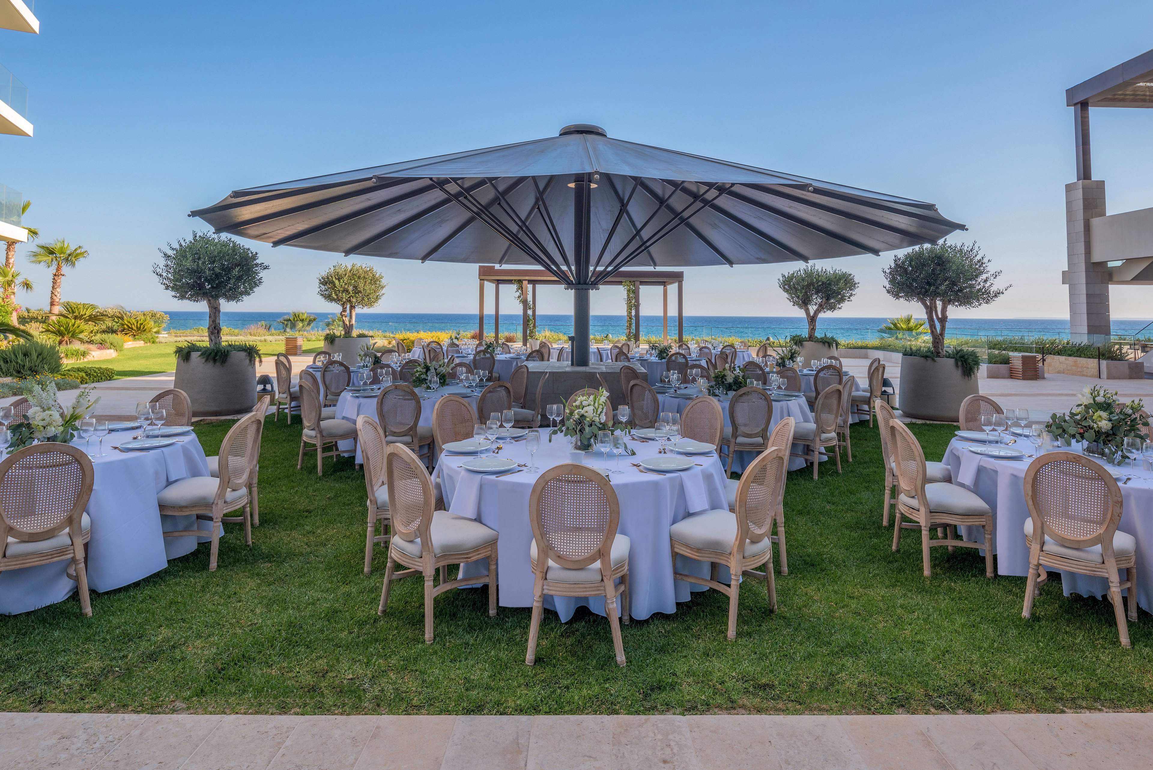 A formal outdoor banquet is arranged on a manicured lawn overlooking the sea. Round tables with white tablecloths and classic cane-back chairs are neatly organized beneath a large central umbrella. Floral centerpieces and glassware decorate each table, suggesting a wedding reception or upscale celebration. Modern resort buildings and palm trees frame the scene under a clear blue sky.