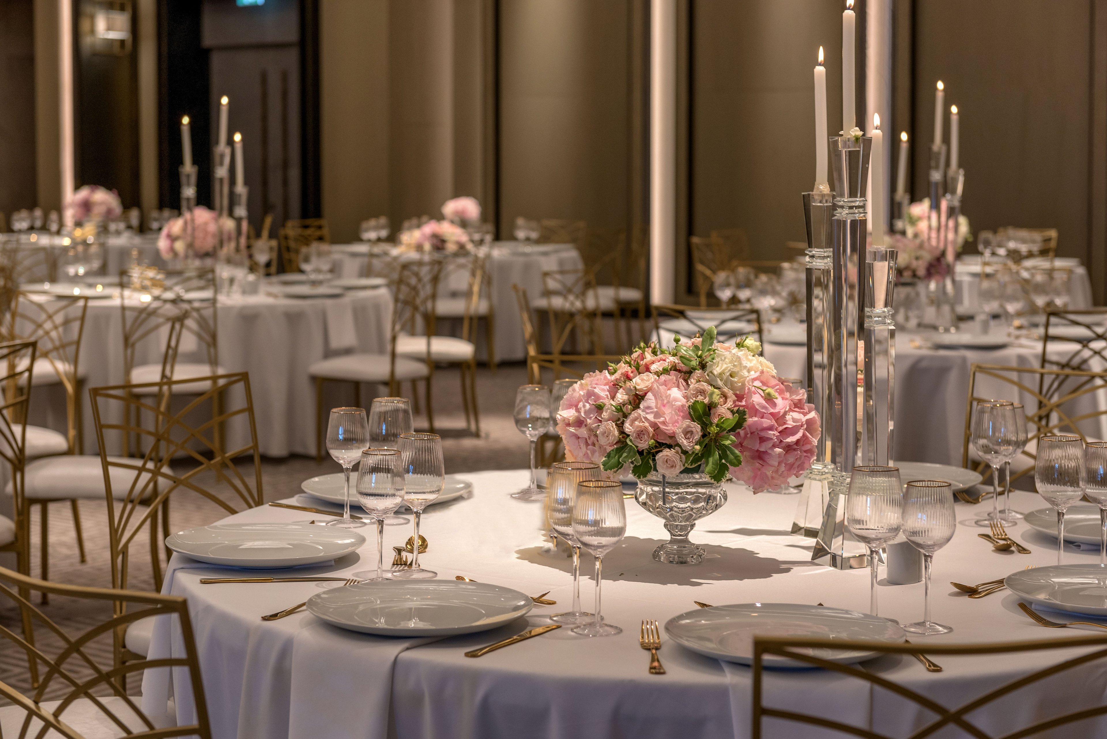 A formal banquet hall is set with round tables covered in white linens. Each table features a large glass vase centerpiece filled with pink and white flowers and greenery. Tall glass candlesticks with white taper candles add height and sophistication to the arrangement. Gold chairs and soft, warm lighting create a luxurious and romantic atmosphere suitable for weddings or upscale events.