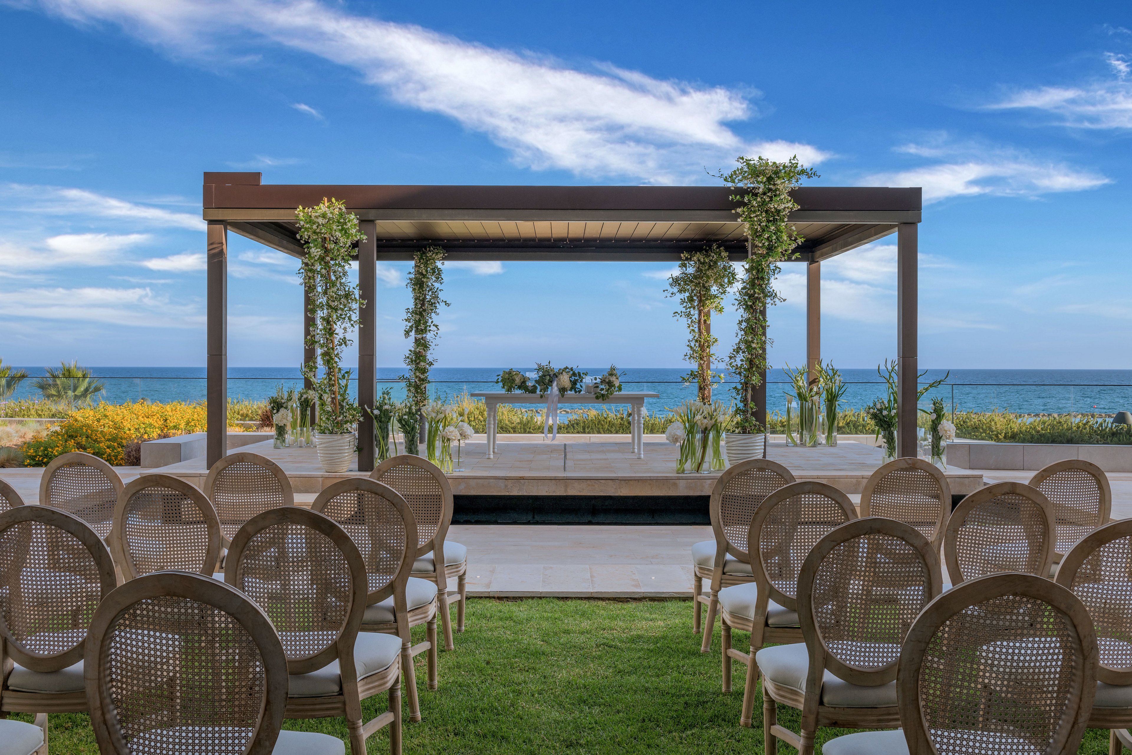 An elegant outdoor wedding ceremony setup is arranged on a lawn facing the ocean. Rows of light-colored chairs are aligned toward a wooden pergola decorated with green vines and floral arrangements. The pergola stands beside a narrow reflecting pool, with the sea and blue sky creating a serene backdrop. Bright flowers and coastal plants frame the scene, enhancing the romantic atmosphere.