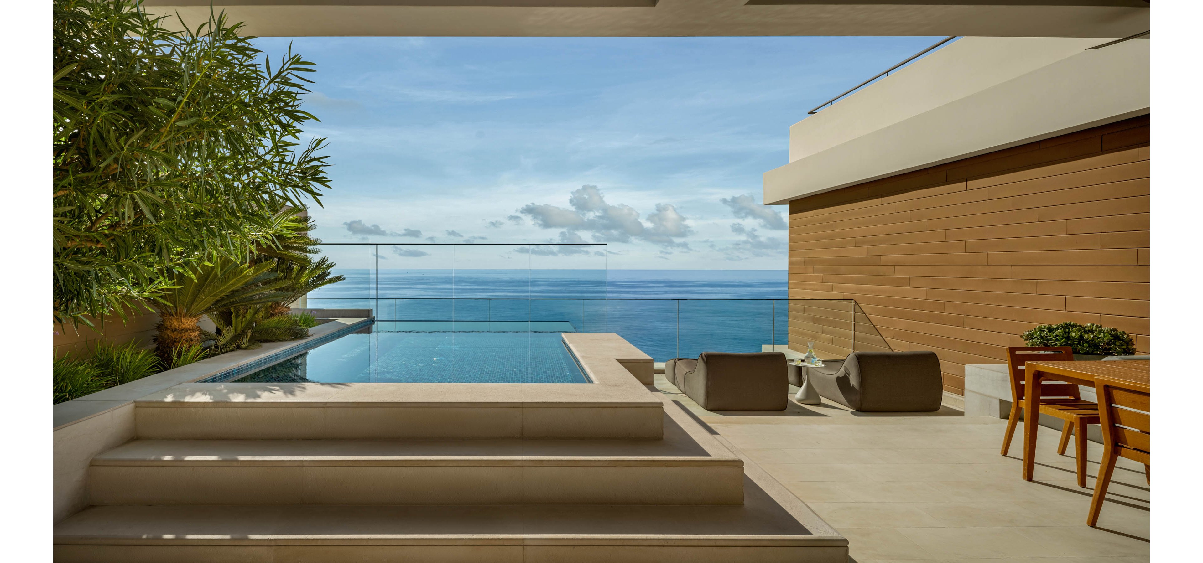 A contemporary terrace overlooks an expansive ocean view with a sleek infinity pool in the foreground. Minimalist lounge chairs and a wooden dining table with chairs create a relaxed outdoor living area. Glass railings frame the horizon, while warm wood paneling contrasts with light stone flooring. Lush greenery softens the architecture, enhancing the tranquil coastal atmosphere.