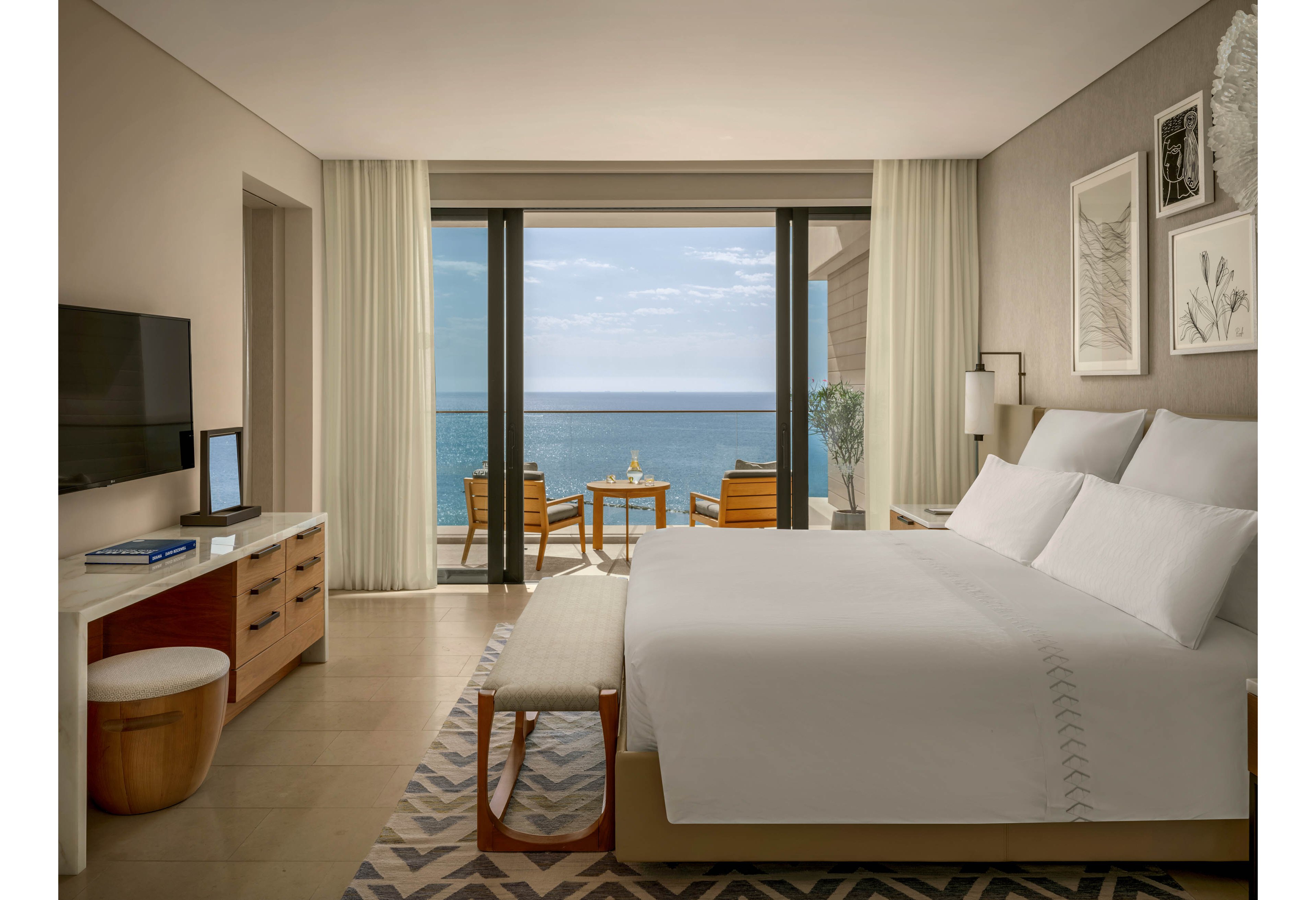 A contemporary hotel bedroom features a large neatly made bed facing a balcony with an expansive ocean view. Neutral-toned walls, light curtains, and minimalist furniture create a calm, upscale atmosphere. A desk with a television, books, and decor sits opposite the bed, while two chairs and a small table are arranged on the balcony. Soft natural light fills the room, highlighting the clean lines and coastal setting.