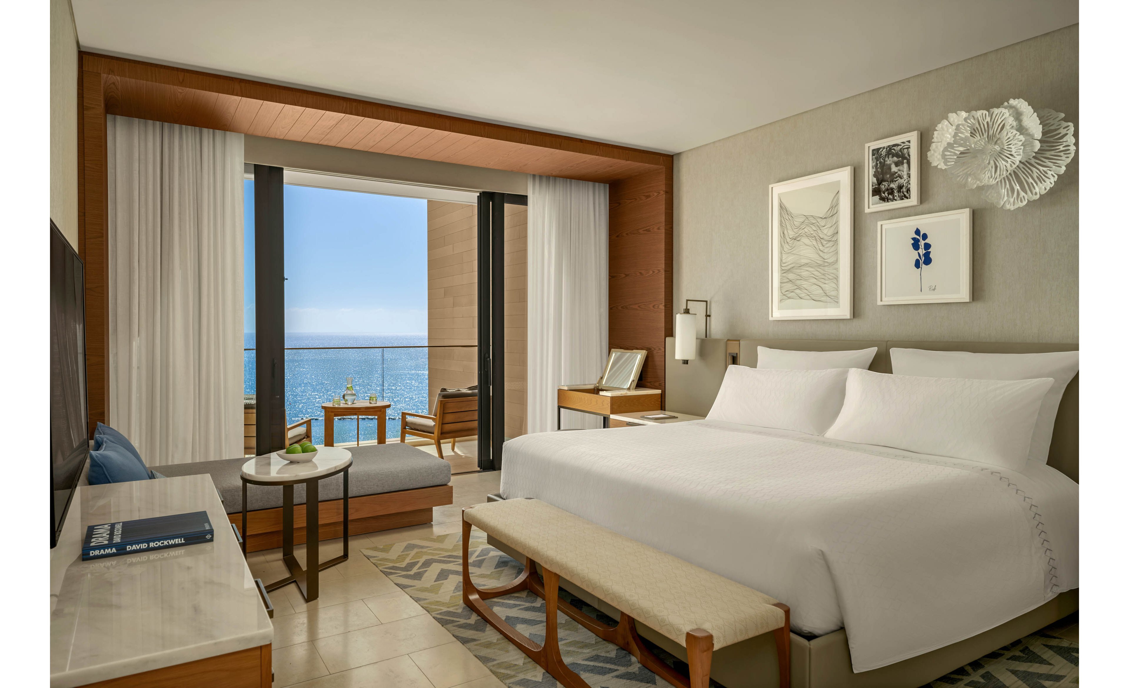 A contemporary hotel room features a large neatly made bed with white linens and a cushioned bench at the foot. The space includes a small seating area with a daybed, side table, and a desk near the balcony. Floor-to-ceiling glass doors open to a balcony with wooden chairs and a table overlooking a calm ocean. Neutral tones, minimalist artwork, and soft lighting create a serene, upscale atmosphere ideal for relaxation.