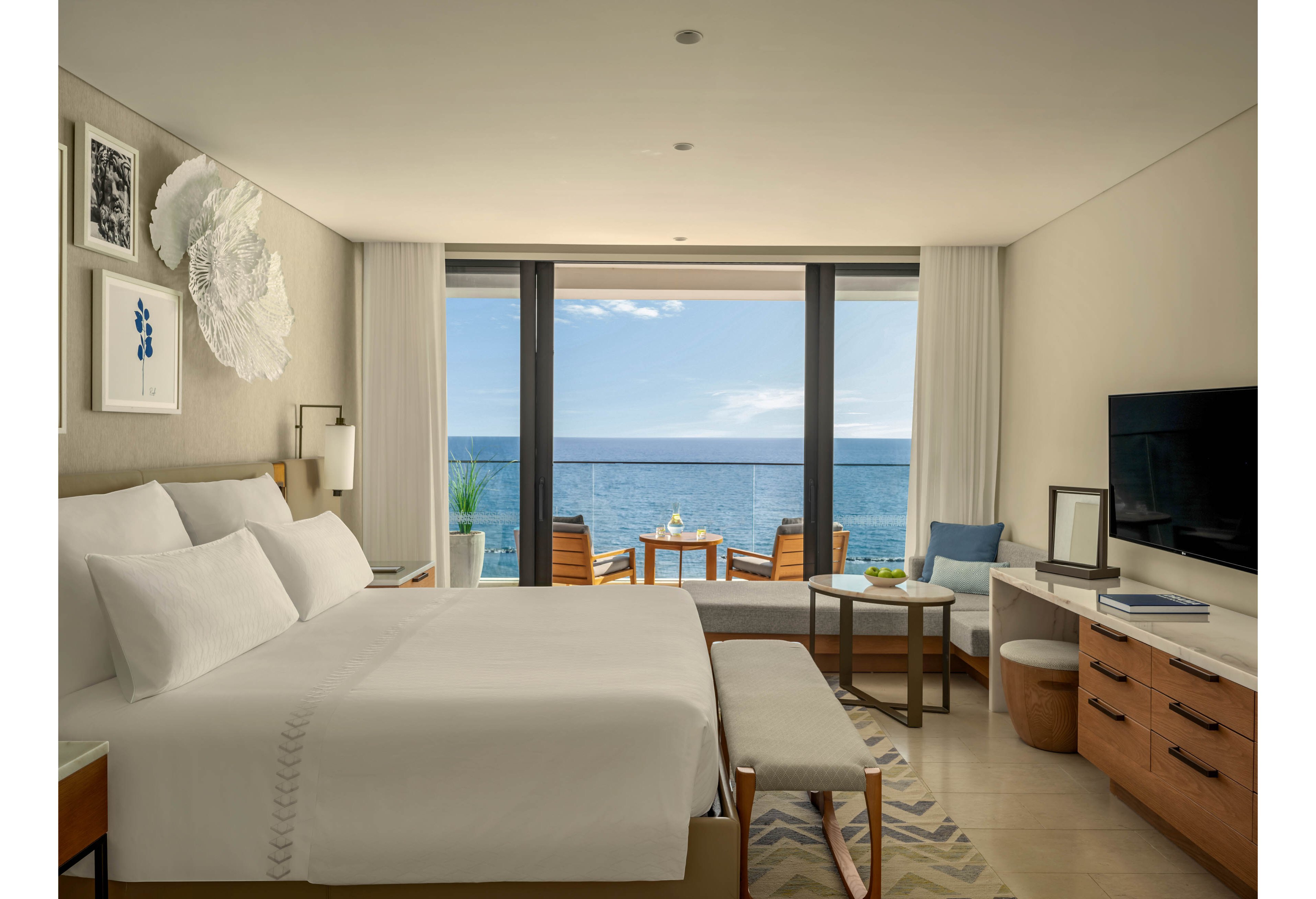 A contemporary hotel room features a large neatly made bed facing a flat-screen television and wooden cabinetry. Floor-to-ceiling glass doors open onto a balcony with chairs and a small table overlooking a calm ocean. Neutral tones, soft lighting, and minimalist artwork create a serene, upscale atmosphere. The space feels airy and inviting, ideal for relaxation or a seaside vacation stay.