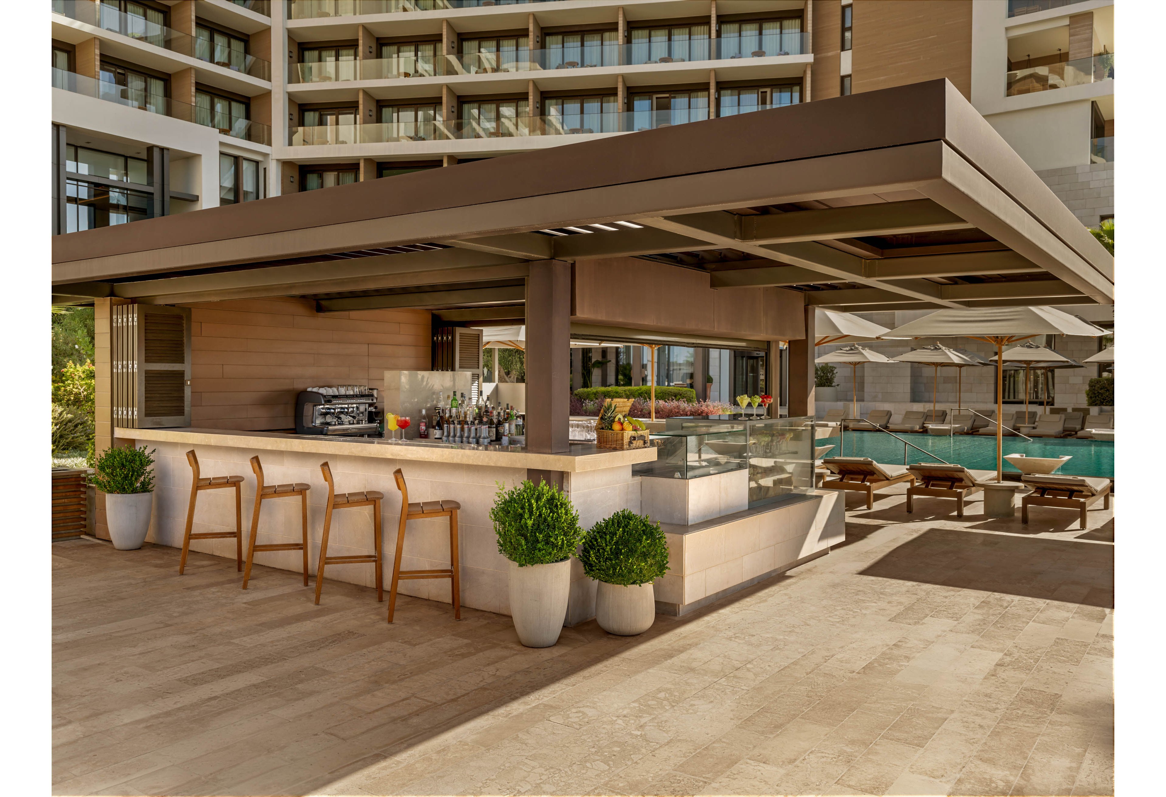 An open-air poolside bar stands in front of a modern resort building. Wooden bar stools line the counter, which is accented by potted green plants and a sleek overhanging roof. Behind the bar, bottles and equipment are neatly arranged, suggesting a relaxed hospitality setting. In the background, sun loungers and umbrellas surround a turquoise swimming pool, creating a calm vacation atmosphere.