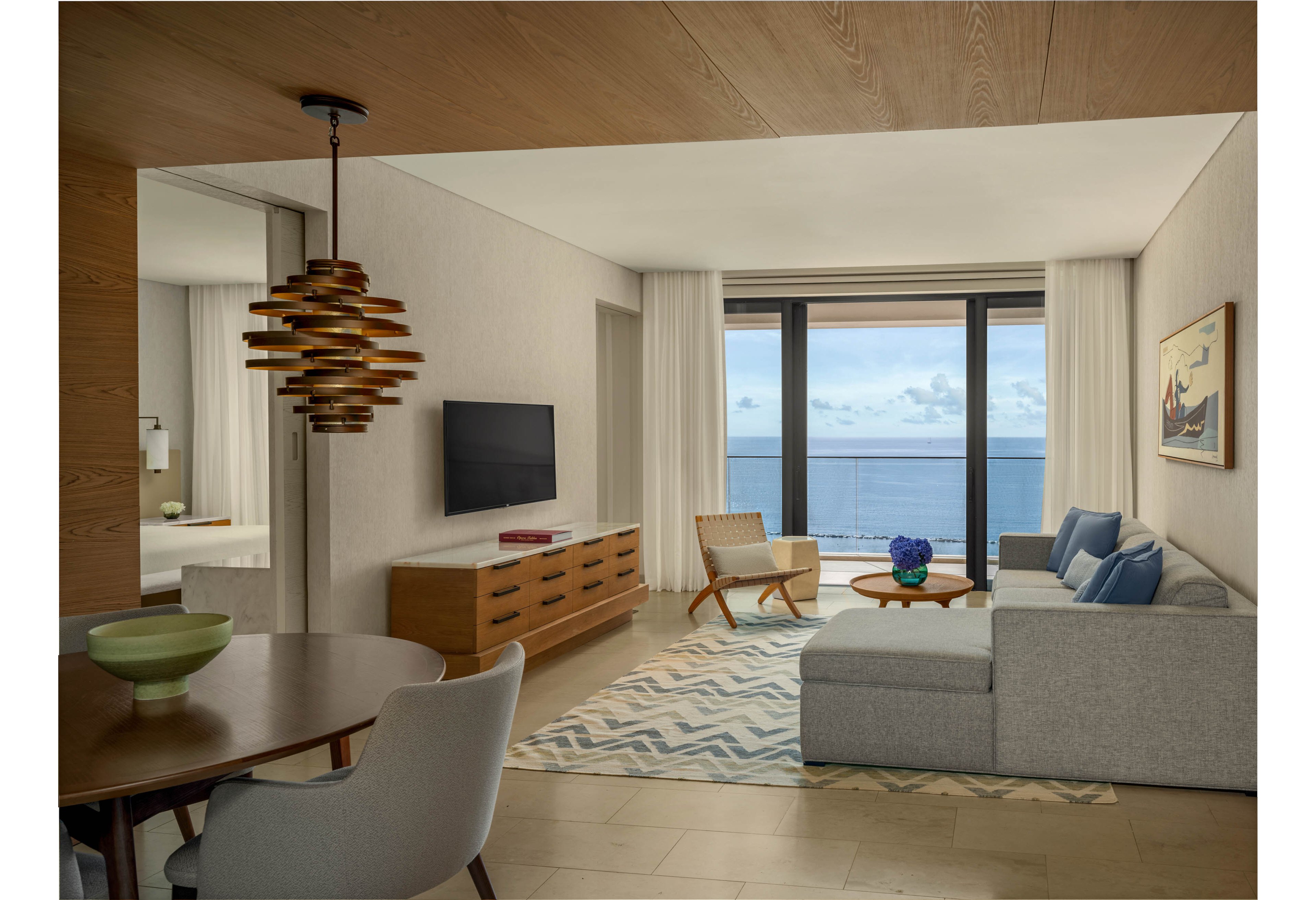 A contemporary hotel suite living room features a light gray sectional sofa facing a wall-mounted television and wooden media console. Large floor-to-ceiling windows reveal a calm ocean view under a cloudy sky, filling the space with natural light. A patterned rug, round wooden coffee table, and accent chair add warmth and texture to the minimalist decor. In the foreground, a dining table with a sculptural pendant light and ceramic bowl enhances the modern coastal ambiance.