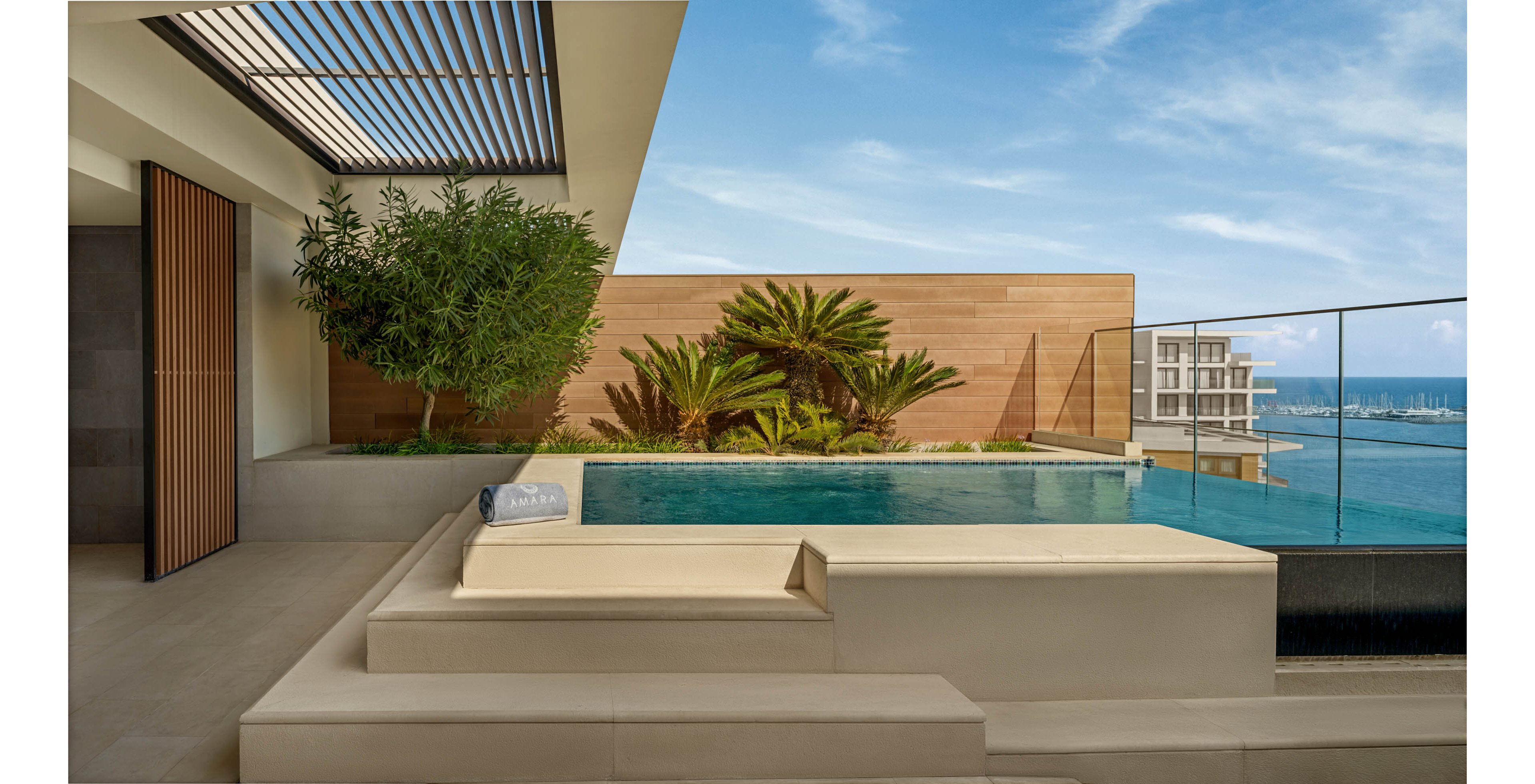 A contemporary rooftop terrace features a sleek infinity pool overlooking a calm ocean. Minimalist beige steps and seating lead up to the water, framed by a glass railing. Lush green plants and a small tree soften the clean architectural lines and wooden privacy wall. In the distance, modern waterfront buildings rise against a clear blue sky, enhancing the luxurious resort atmosphere.
