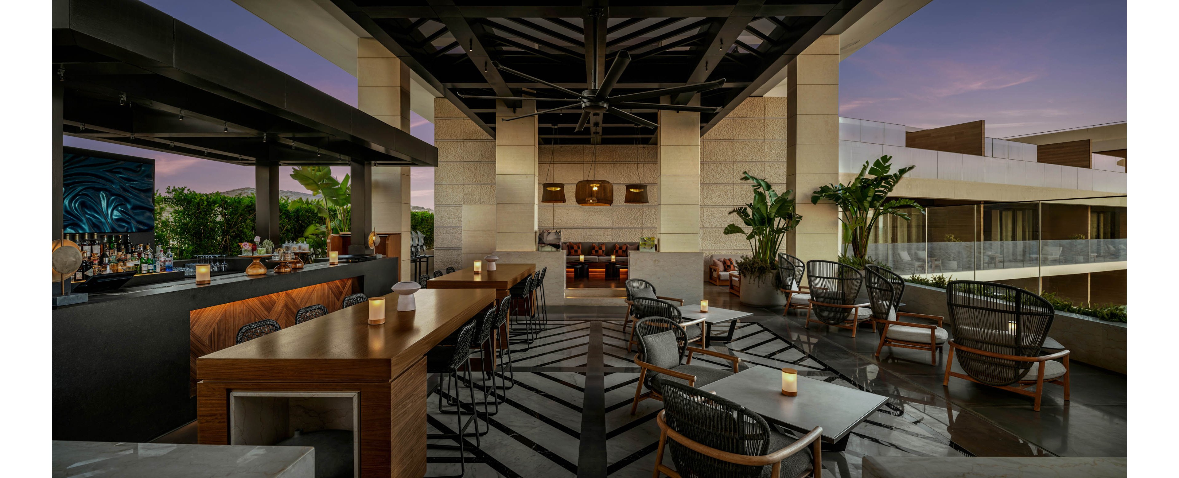 A contemporary rooftop bar and lounge features sleek wooden tables and cushioned chairs arranged under a partially open ceiling structure. The setting overlooks a multi-level building with glass railings and lush greenery, creating an upscale urban atmosphere. Warm ambient lighting from candles and recessed fixtures contrasts with the cool tones of the evening sky. The space appears ready for guests, emphasizing relaxation, socializing, and stylish outdoor dining.