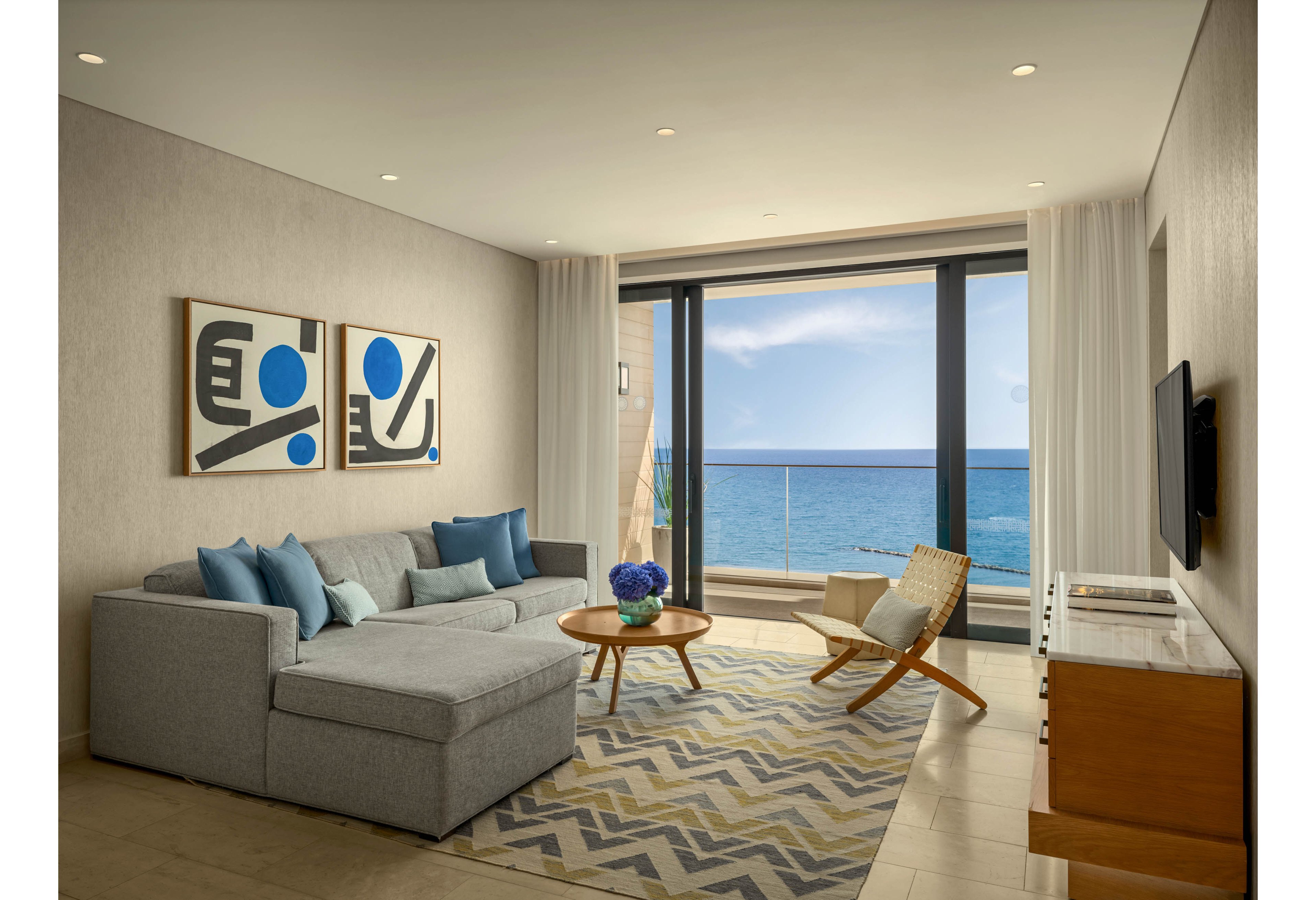A contemporary living room features a large gray sectional sofa accented with blue pillows and abstract artwork on the wall. Floor-to-ceiling glass doors open to a balcony overlooking a bright blue ocean. A wooden rocking chair, round coffee table with flowers, and zigzag patterned rug add warmth and texture to the neutral interior. Recessed ceiling lights and natural daylight create a calm, inviting atmosphere.
