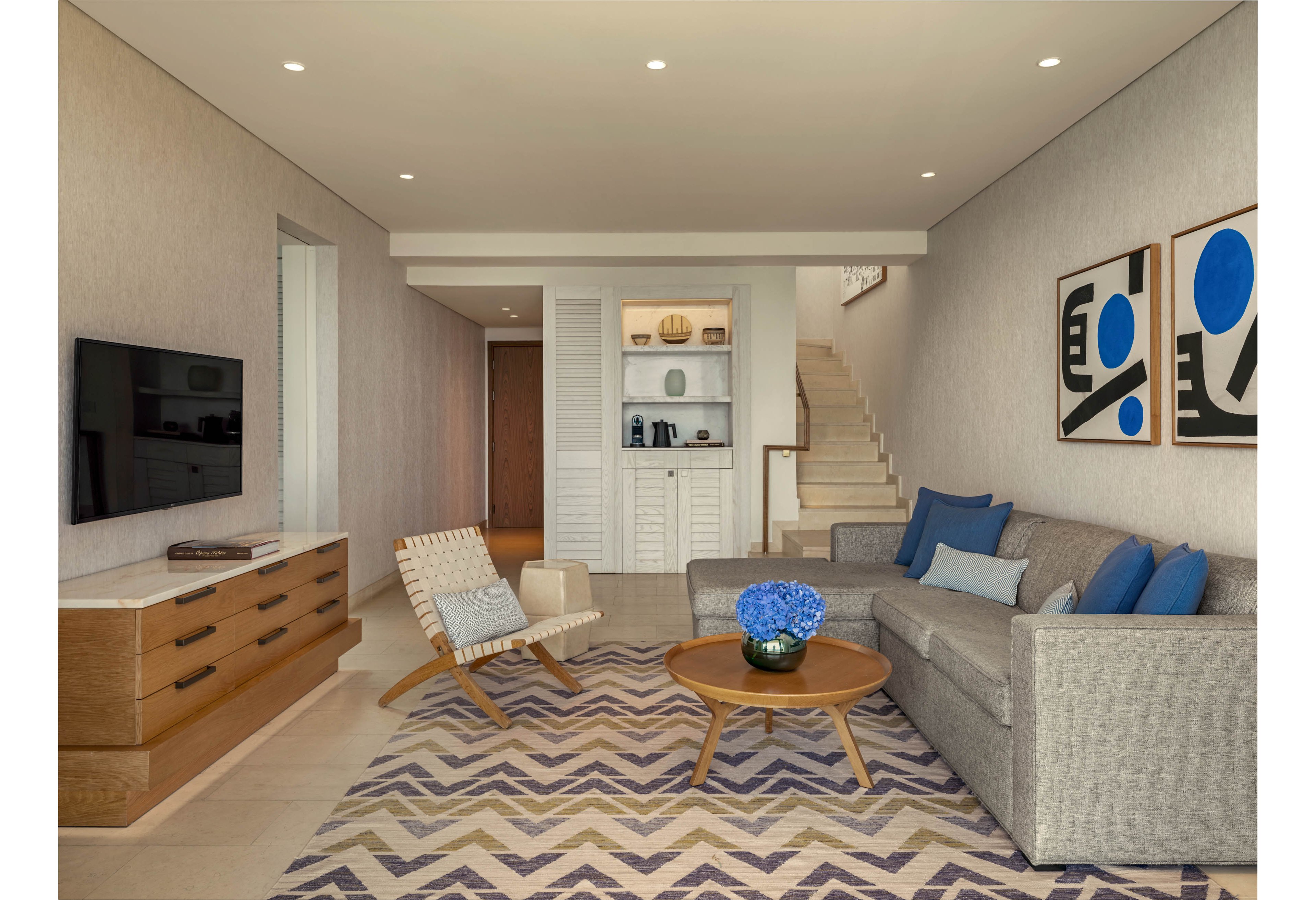 A contemporary living room features a gray sofa, wooden coffee table, and patterned rug in a neutral-toned interior. Blue accent pillows and a vase of blue flowers add color to the space, complemented by abstract artwork on the wall. A flat-screen television sits above a wooden media console, while a woven lounge chair and side table complete the seating area. In the background, a staircase and built-in shelving with decorative objects lead to another level, creating a clean and organized atmosphere.